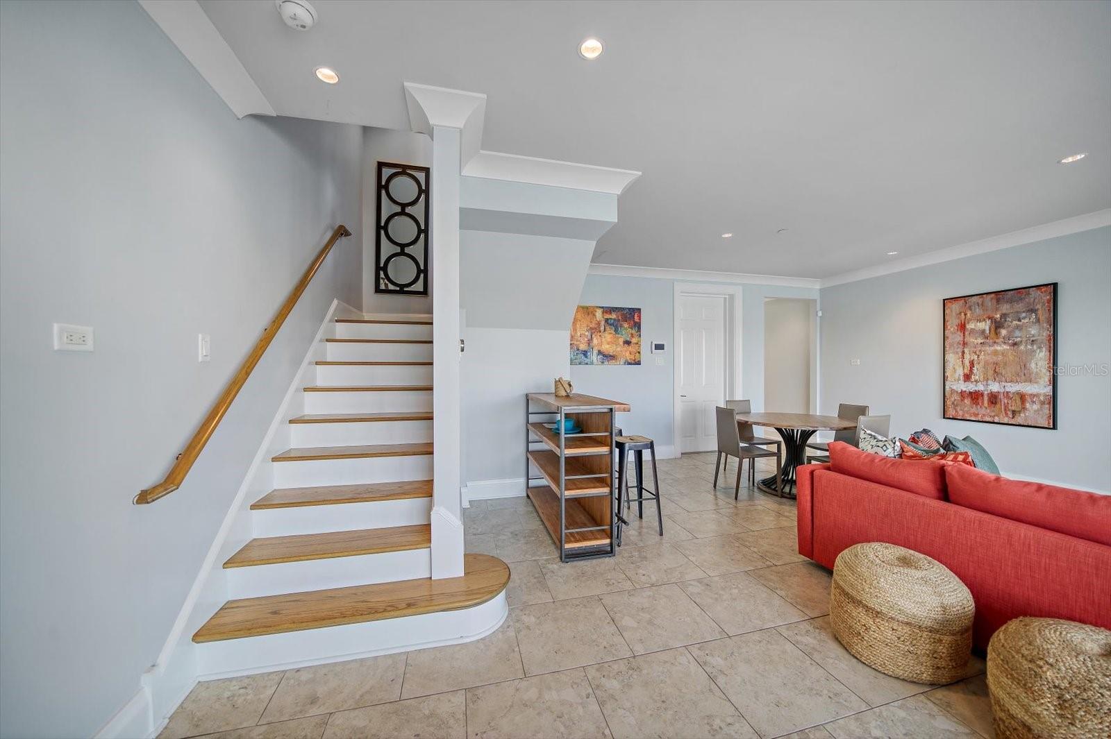 The lower-level includes a two-car garage and a spacious foyer/media room featuring a mounted TV and floor-to-ceiling multi-panel sliding glass doors that create a dramatic wall of glass, instantly framing tranquil water views.