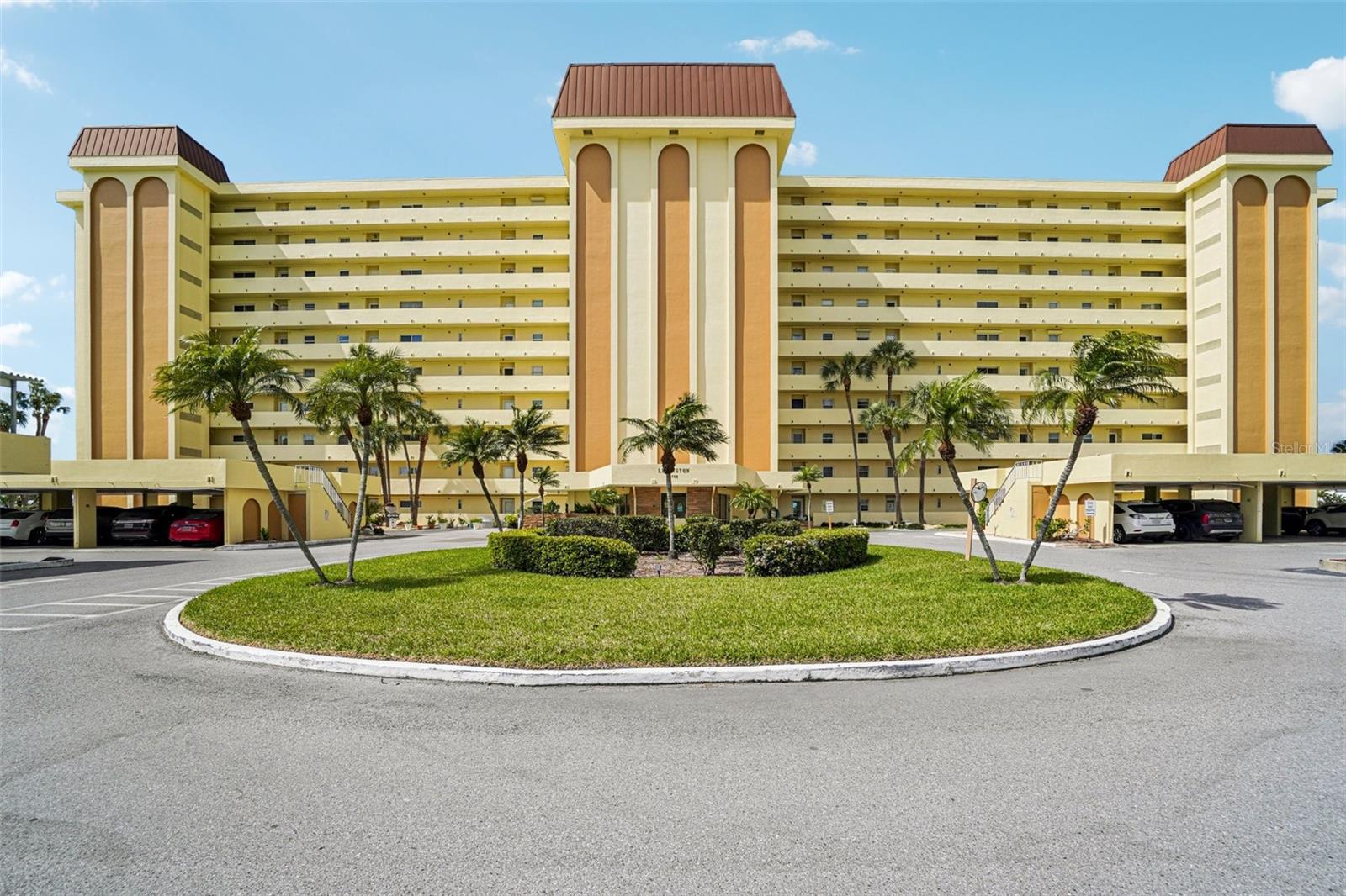 Building Exterior / Community Entry – This well-maintained mid-rise community makes a striking first impression with its bold architectural design, covered parking, and lushly landscaped circular entry featuring mature palm trees and manicured hedges all set against a bright Florida sky.