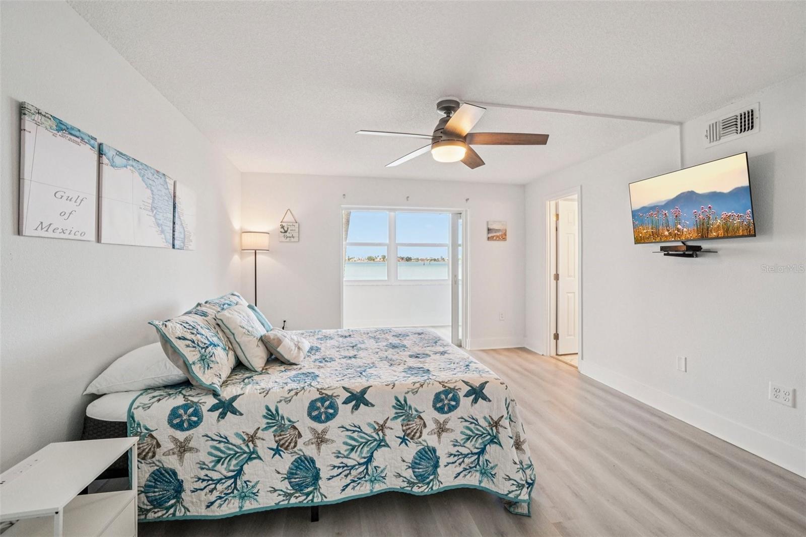 Primary Bedroom with Water View – Wake up to breathtaking water views from this serene primary bedroom, which features direct access to an en suite bathroom, a wall-mounted TV, a modern ceiling fan, and light LVP flooring — a true coastal retreat within your own home.