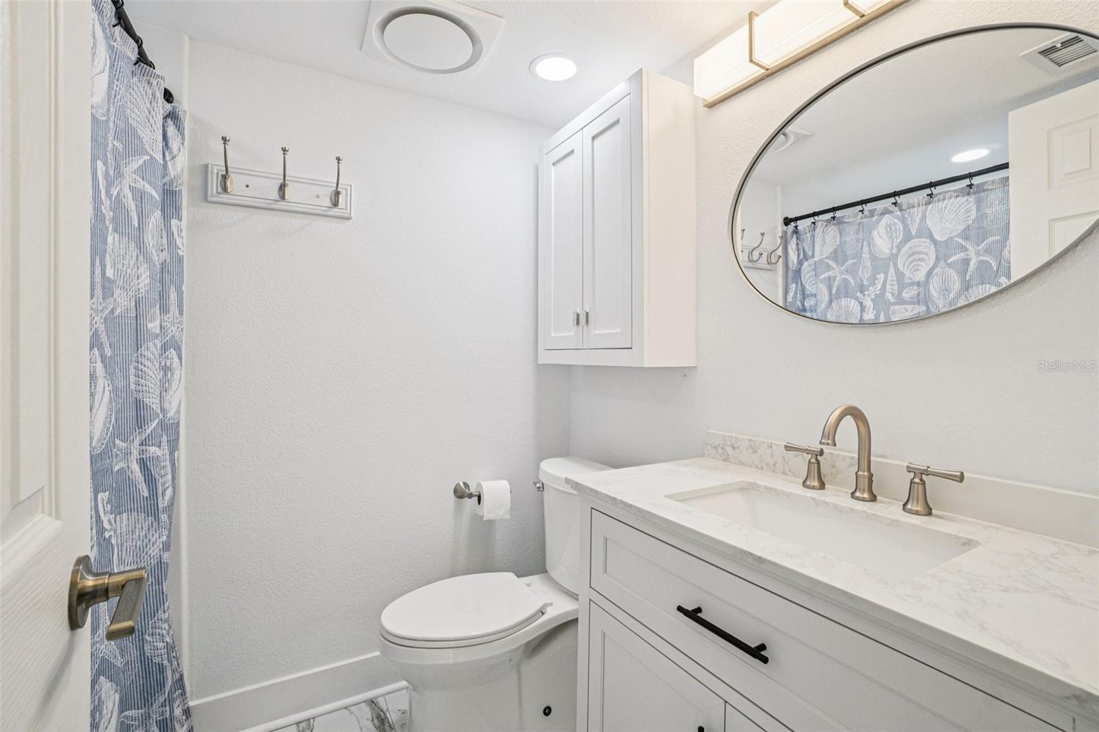 Primary En Suite Bathroom – The primary en suite bathroom features a white shaker vanity with quartz countertop, brushed nickel fixtures, an elegant oval mirror with lighted vanity bar, upper cabinet storage, and a coastal-themed tub/shower combo — a beautifully finished private retreat off the main bedroom.