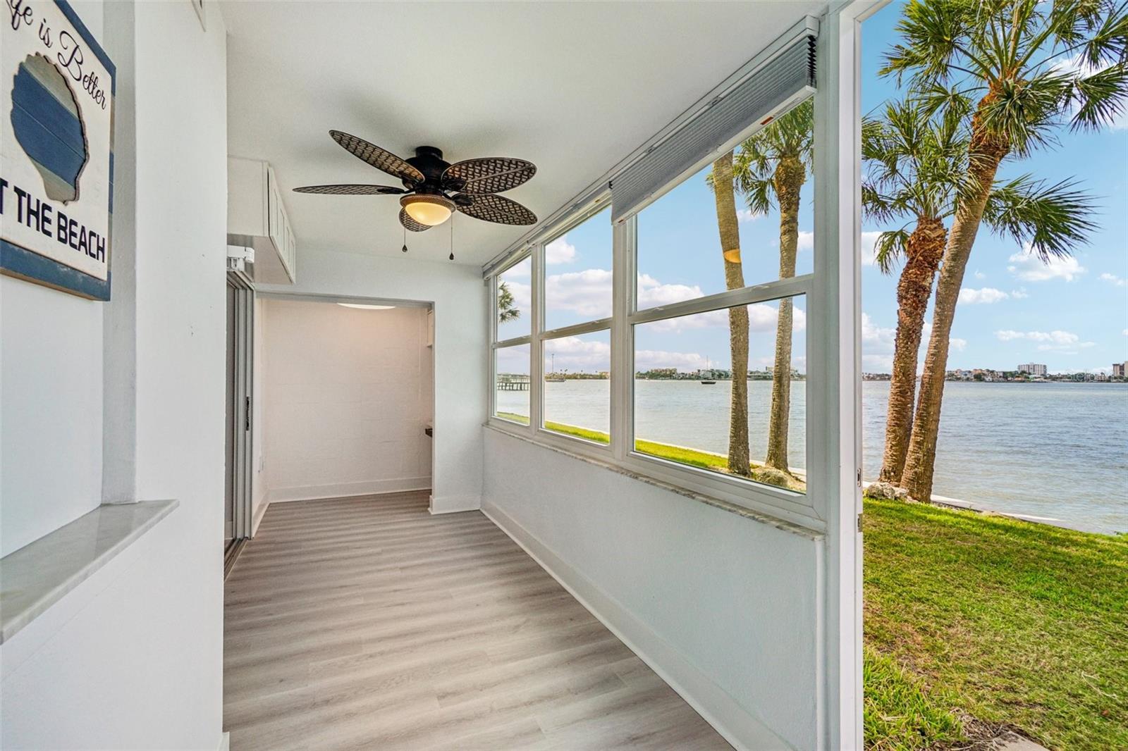 Florida Room / Waterfront Sunroom – This stunning enclosed Florida room is an absolute showstopper — floor-to-ceiling windows frame an unobstructed panoramic water view lined with swaying palm trees, while a tropical-style ceiling fan and light LVP flooring create a breezy, resort-like atmosphere perfect for morning coffee or evening sunsets.