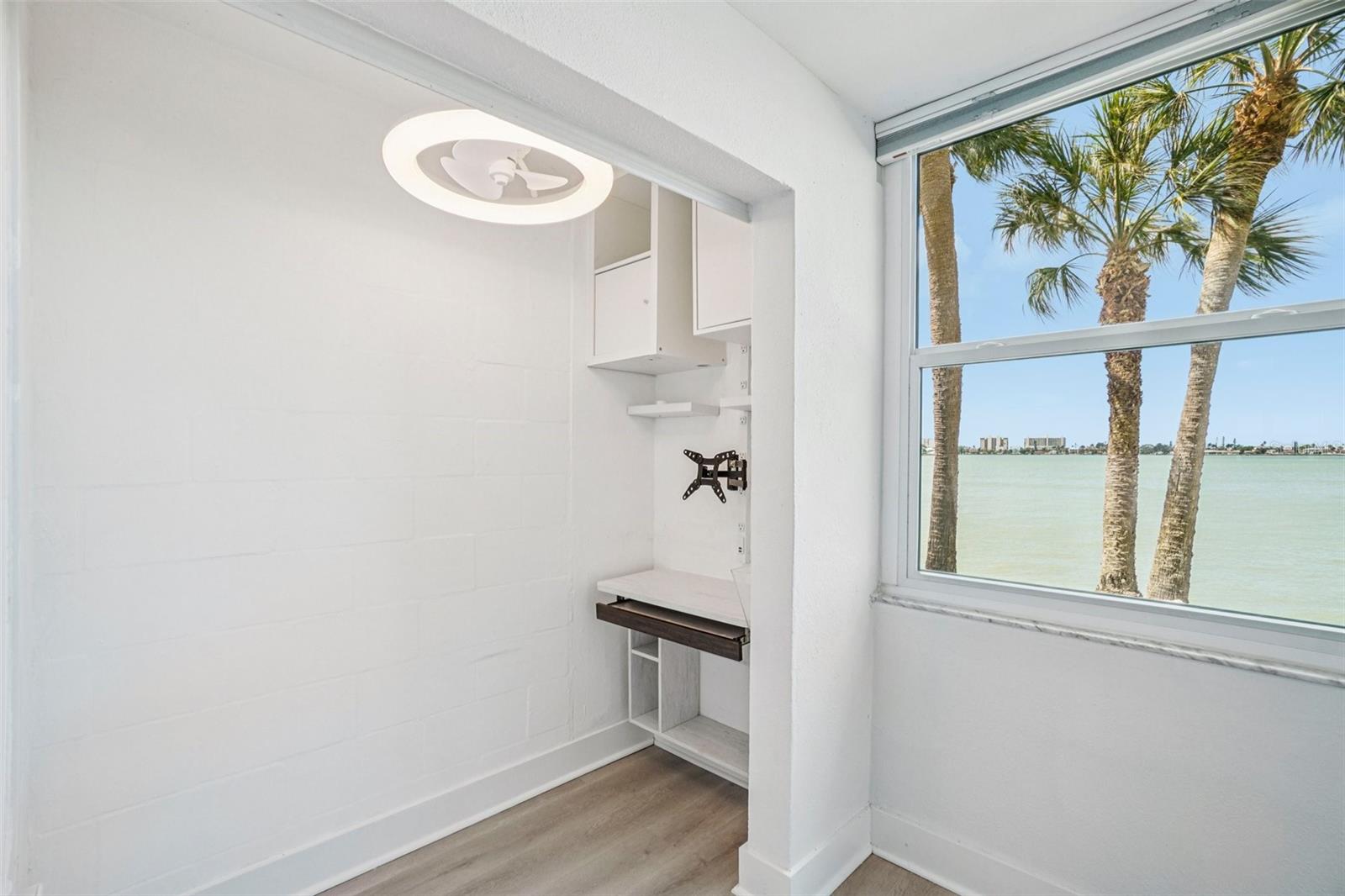 Home Office / Workstation Nook – Tucked just off the living room, this clever built-in workstation features upper cabinetry, open shelving, a fold-down desk surface, and a TV mount — all positioned directly beside a large water-view window, making it one of the most inspired home office setups imaginable.