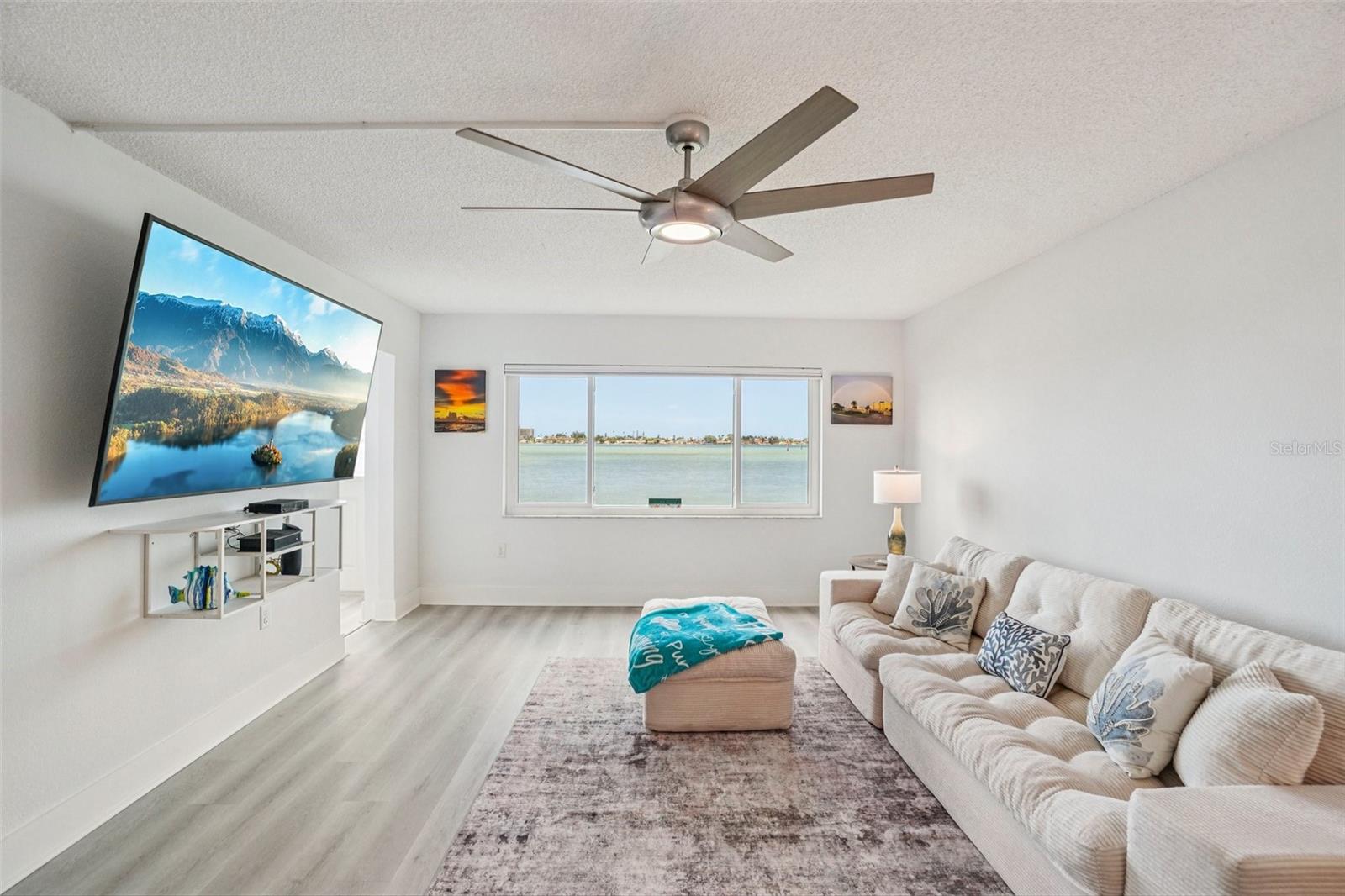 Living Room with Water ViewThe sunlit living room offers a stunning water view framed by a large picture window, complemented by a modern oversized ceiling fan, light LVP flooring, and a coastal-inspired design that brings the outdoors in.