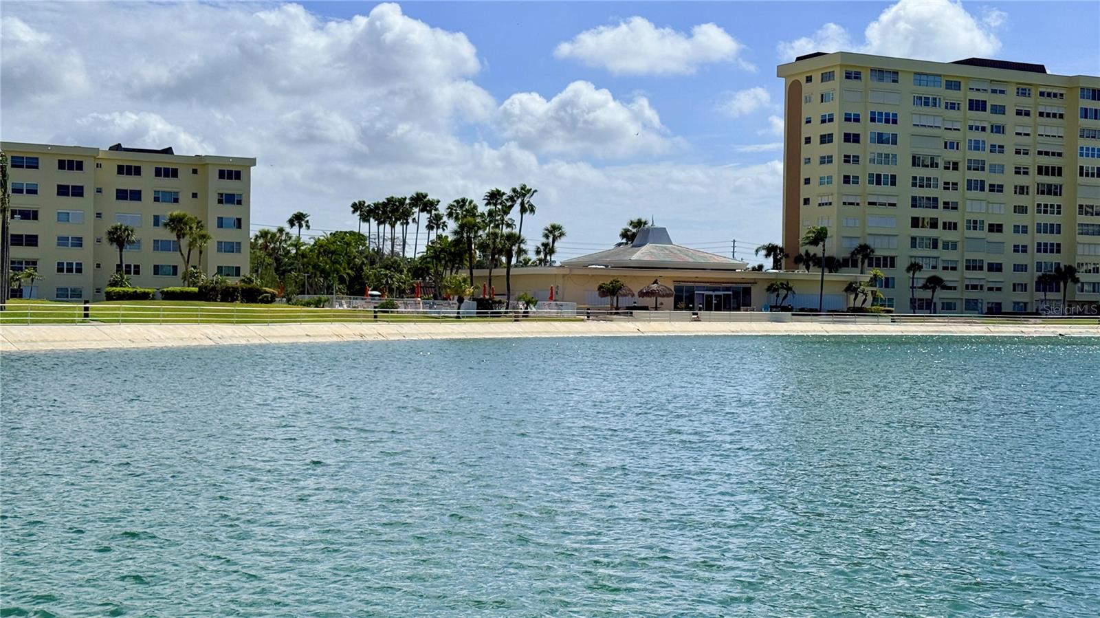 Community Clubhouse & Pool (Water View) – This stunning view from the water showcases the community's expansive clubhouse and second pool area, flanked by towering palm trees and manicured waterfront grounds — a resort-style amenity campus that perfectly captures the laid-back Florida waterfront lifestyle.