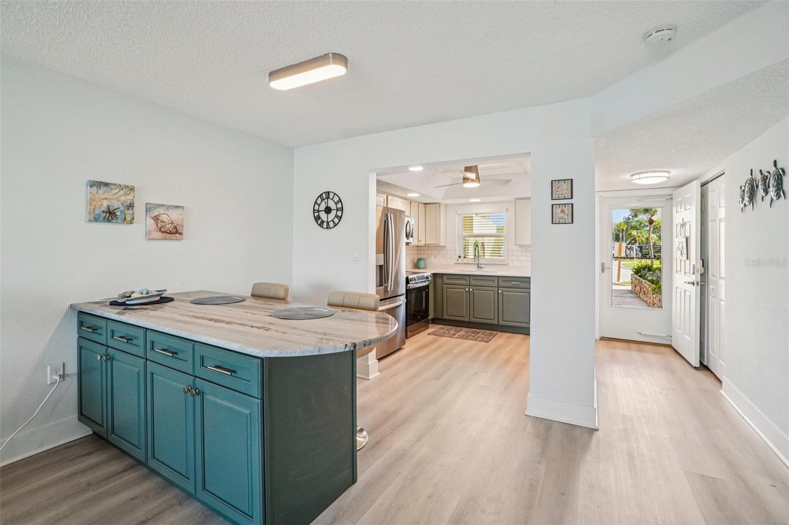Dining Area & Kitchen Overview. This bright, open layout connects a stylish teal-cabinet dining island with a stone countertop to the updated kitchen beyond, featuring stainless steel appliances, subway tile backsplash, and light luxury vinyl plank flooring throughout — a seamless flow perfect for casual dining and entertaining.
