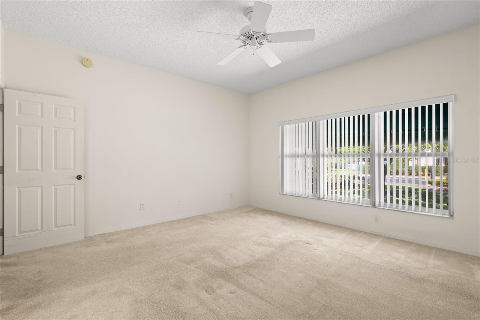 Primary bedroom featuring a wide window with vertical blinds, bringing in natural light and a pleasant view of the surrounding greenery.