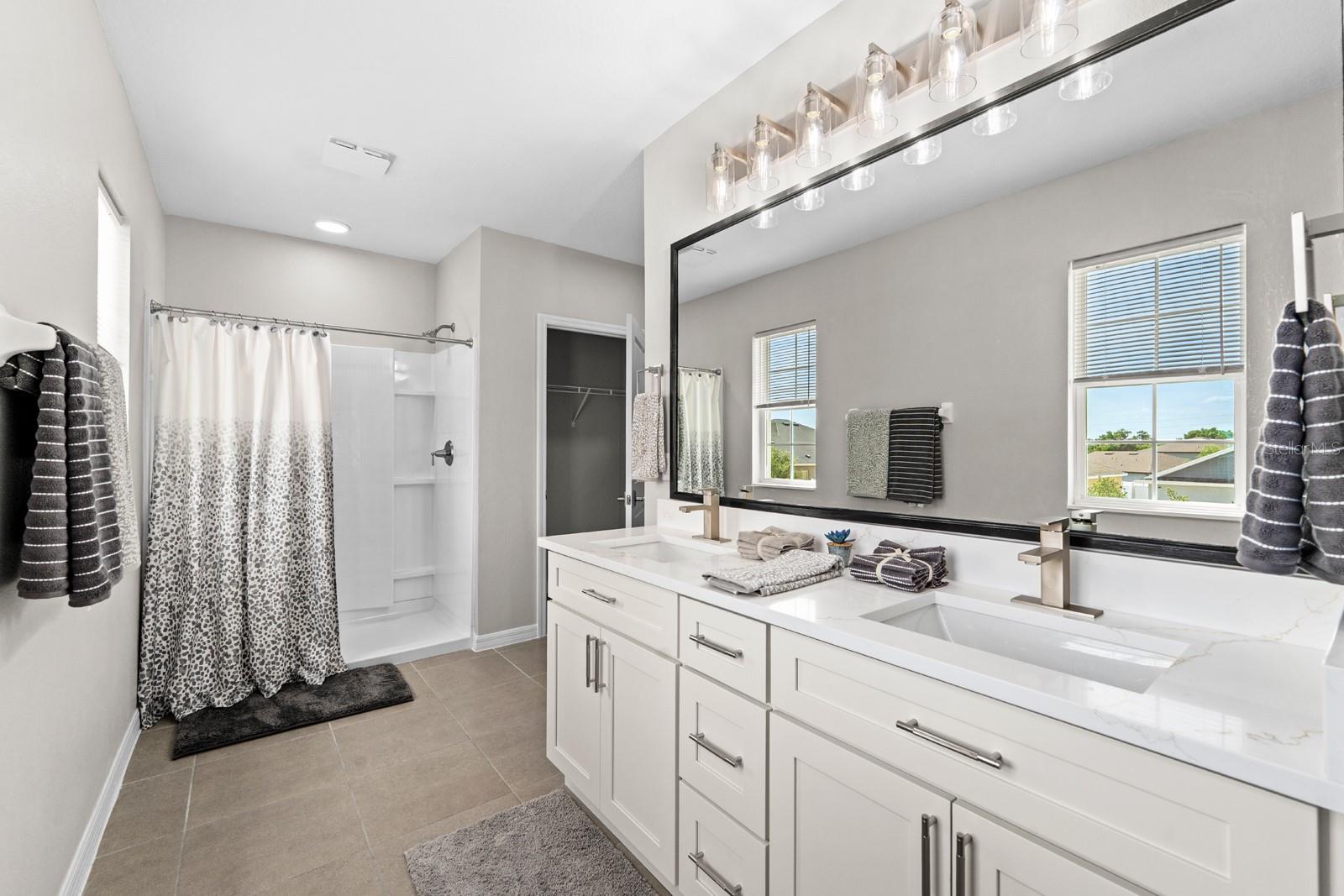The Primary En Suite features a new double-sink vanity with modern faucets, a new mirror, new lighting, new hardware in the shower, a new toilet, and another smaller walk-in closet.
