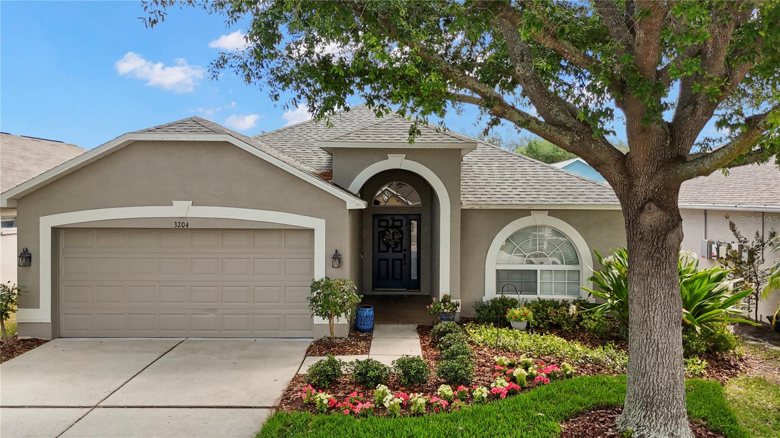Welcome to this charming single-story home featuring a welcoming arched entry, fresh neutral exterior with white trim, a spacious two-car garage, and a beautifully landscaped front yard with vibrant flower beds and mature shade trees under sunny Florida skies.