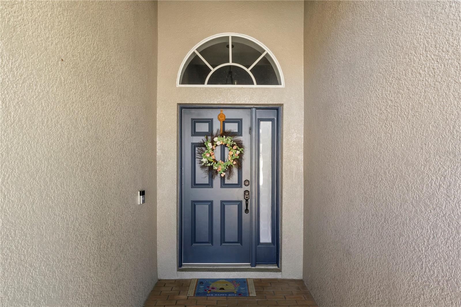 Charming front entry featuring a navy blue double door with sidelight and transom window, accented by a welcoming floral wreath and covered brick porch.