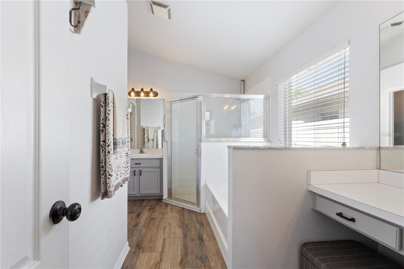 Bright and modern bathroom featuring a glass-enclosed shower, separate soaking tub, gray vanity with white countertop, and a convenient built-in vanity desk with stool, all accented by wood-look flooring and abundant natural light.