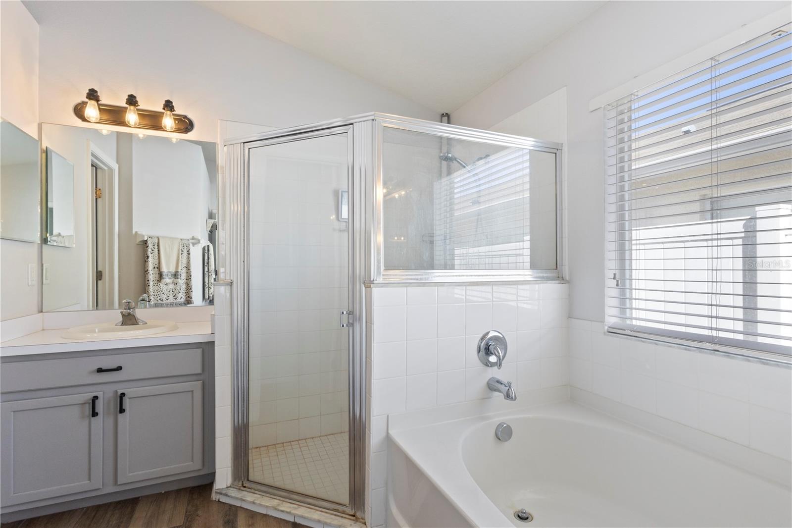 Modern bathroom featuring a gray vanity with white countertop and integrated sink, large glass-enclosed shower, separate soaking tub, and a window providing natural light.