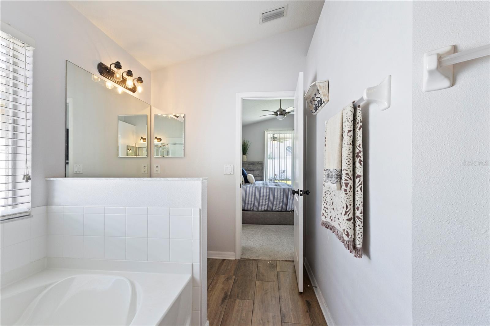 Bright and functional bathroom featuring a soaking tub, white tiled half-wall, large mirror with vanity lighting, wood-look flooring, and a direct view into the adjacent primary bedroom through an open doorway.