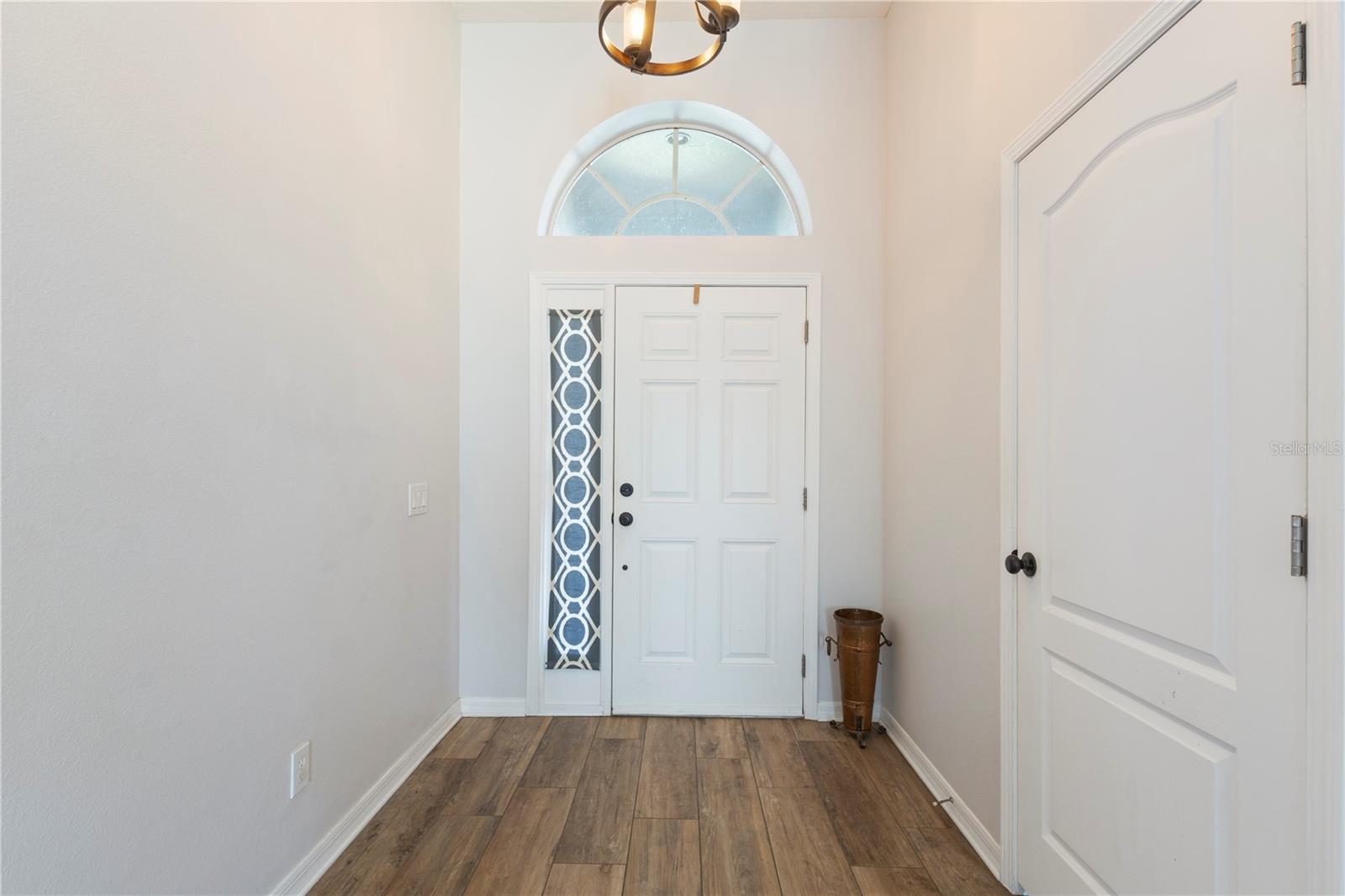 This bright, welcoming entryway features a white six-panel front door with a decorative blue-and-white sidelight, an arched transom window above, light neutral walls, wood-look flooring, and a simple chandelier.