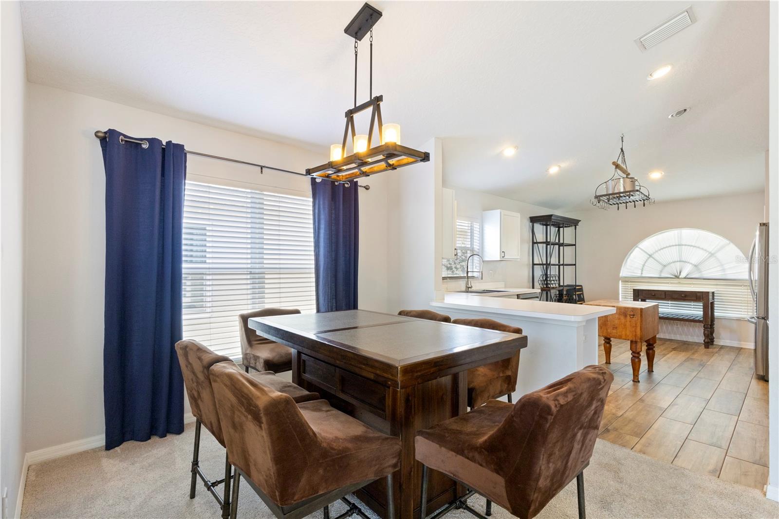 Bright open-concept dining area with stylish chandelier, large window with blue curtains, and seamless flow into a modern kitchen featuring a breakfast bar and abundant natural light.