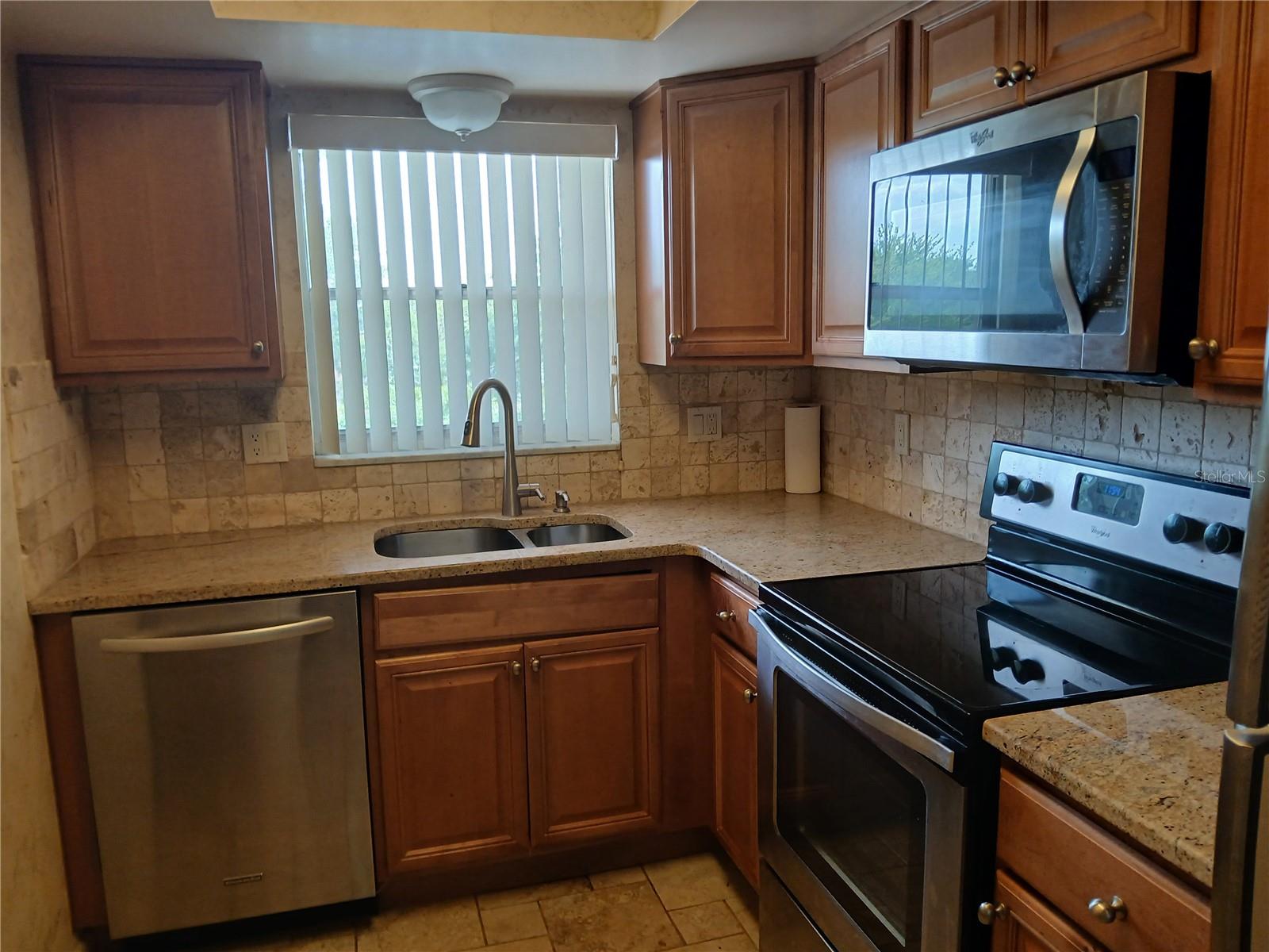 Close up of the kitchen showing the granite counter tops, stainless steel, sink, dishwasher, stove, microwave and the nice window over the sink offering plenty of daylight and a sky view.