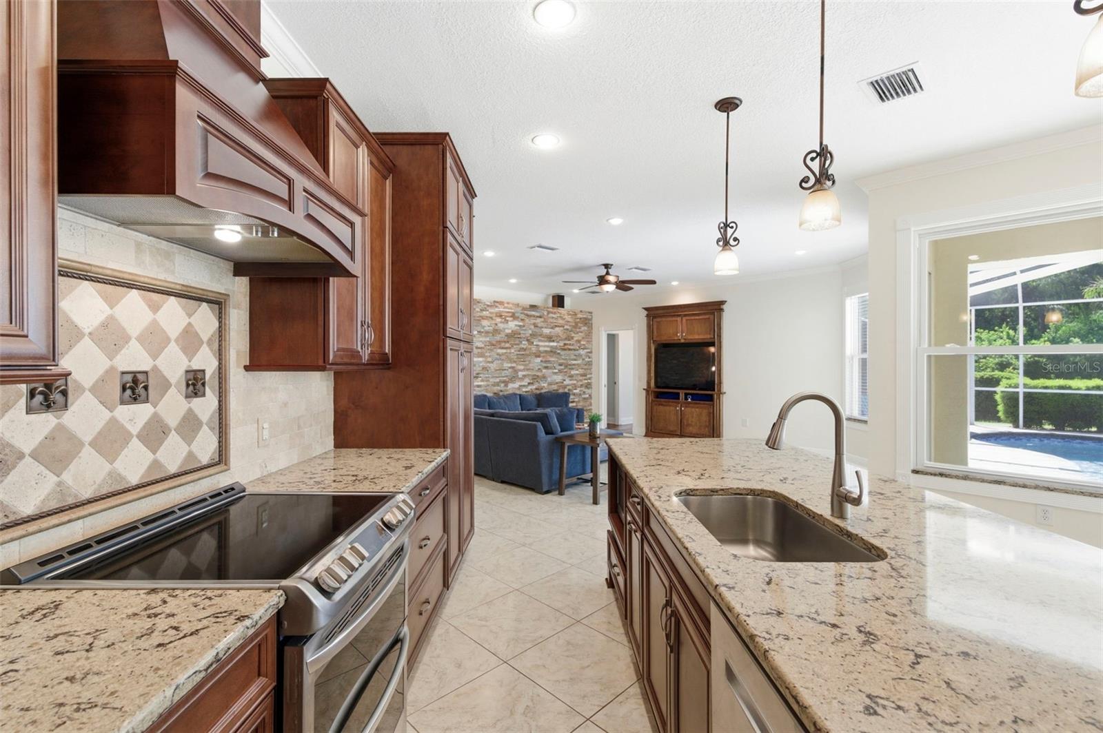 Beautifully appointed kitchen designed for both everyday living and entertaining, featuring rich wood cabinetry, granite countertops, and a large center island with seating. Stainless steel appliances, decorative range hood, and custom tile backsplash add a refined touch, while pendant lighting and recessed lighting create a warm, inviting atmosphere. The oversized island serves as the perfect gathering space for casual dining, hosting, or staying connected with family and guests.