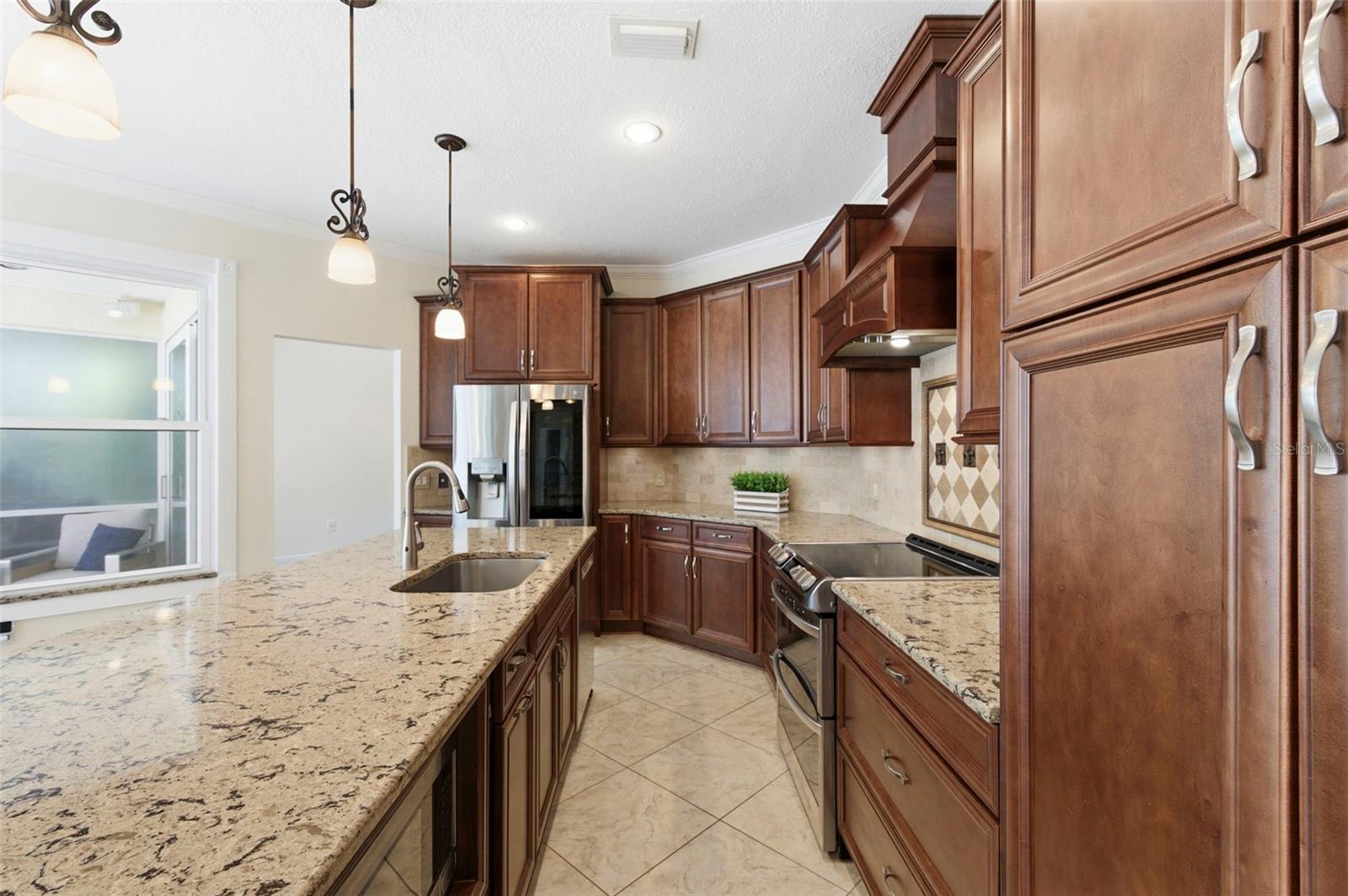 Beautifully appointed kitchen designed for both everyday living and entertaining, featuring rich wood cabinetry, granite countertops, and a large center island with seating. Stainless steel appliances, decorative range hood, and custom tile backsplash add a refined touch, while pendant lighting and recessed lighting create a warm, inviting atmosphere. The oversized island serves as the perfect gathering space for casual dining, hosting, or staying connected with family and guests.