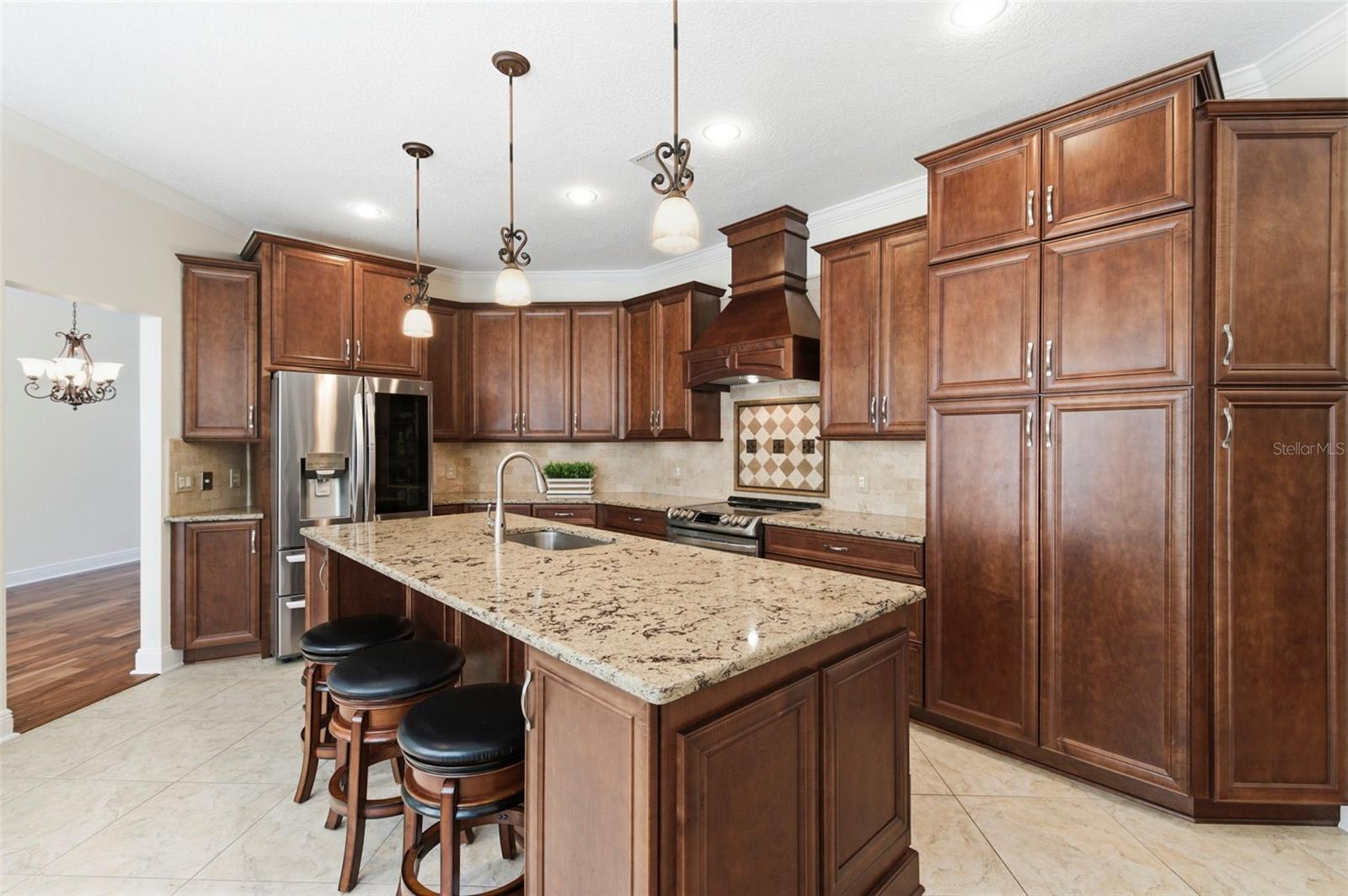 Beautifully appointed kitchen designed for both everyday living and entertaining, featuring rich wood cabinetry, granite countertops, and a large center island with seating. Stainless steel appliances, decorative range hood, and custom tile backsplash add a refined touch, while pendant lighting and recessed lighting create a warm, inviting atmosphere. The oversized island serves as the perfect gathering space for casual dining, hosting, or staying connected with family and guests.