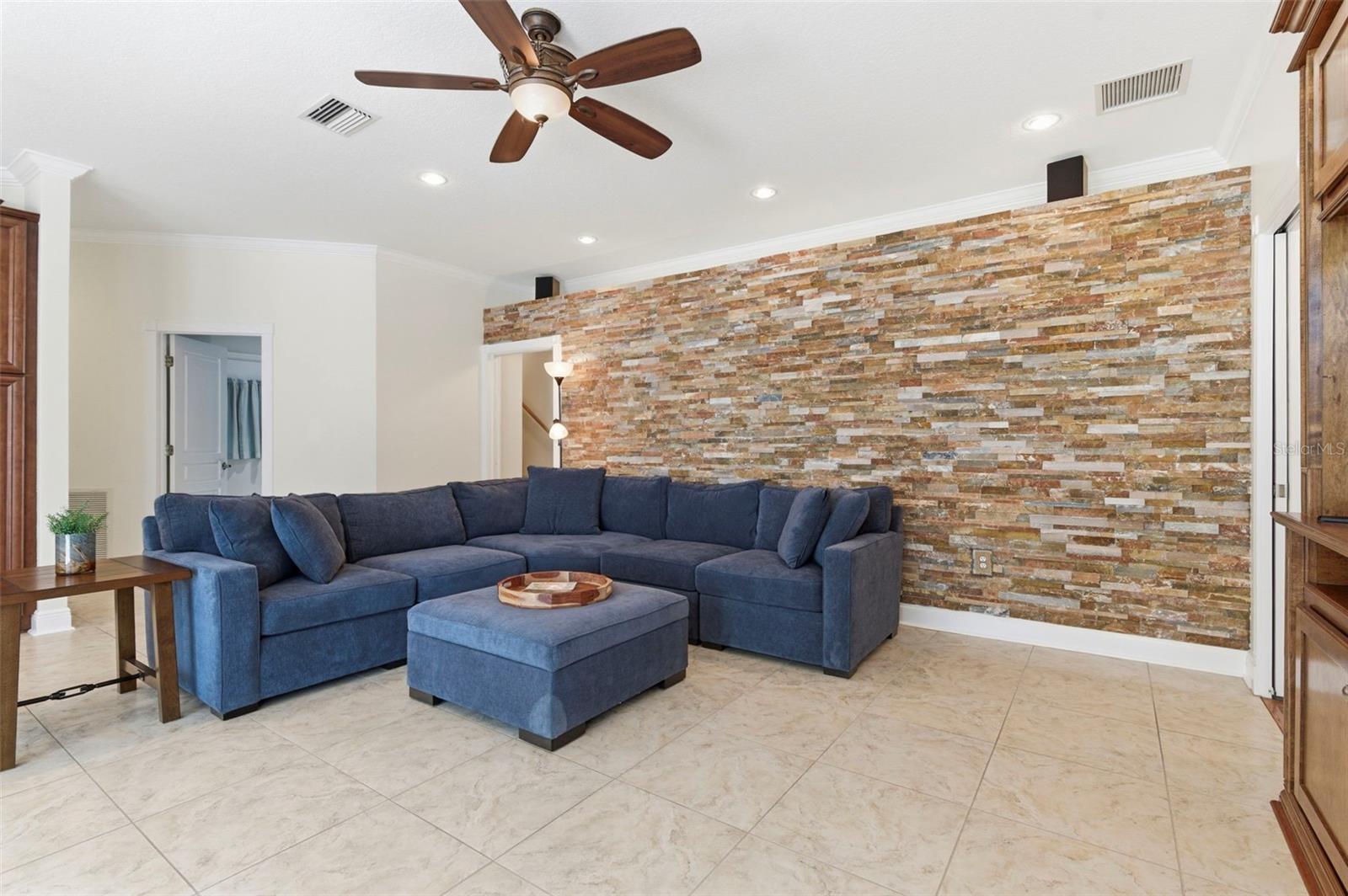 This spacious living area combines comfort and style with an open layout designed for both relaxing and entertaining. A striking stone accent wall creates a warm focal point, complemented by tile flooring, crown molding, and recessed lighting throughout. Natural light pours in through large windows, enhancing the bright and airy feel of the space. The room flows seamlessly into adjacent living areas, making it ideal for gatherings or everyday living.