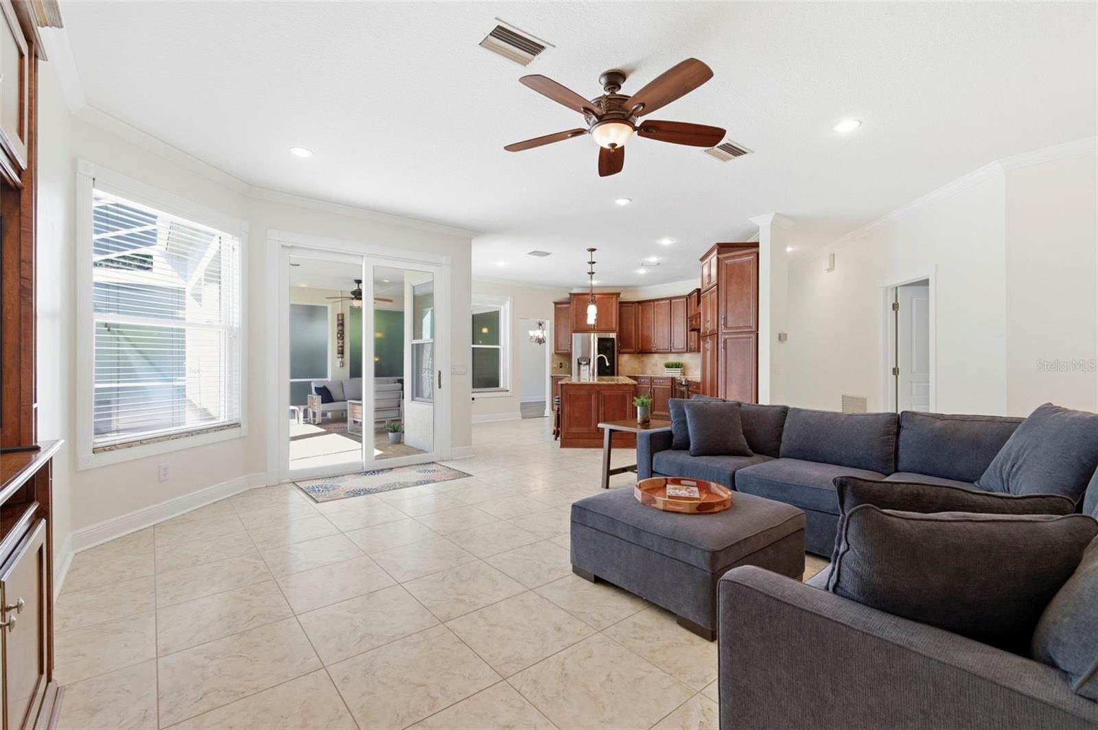 This spacious living area combines comfort and style with an open layout designed for both relaxing and entertaining. A striking stone accent wall creates a warm focal point, complemented by tile flooring, crown molding, and recessed lighting throughout. Natural light pours in through large windows, enhancing the bright and airy feel of the space. The room flows seamlessly into adjacent living areas, making it ideal for gatherings or everyday living.