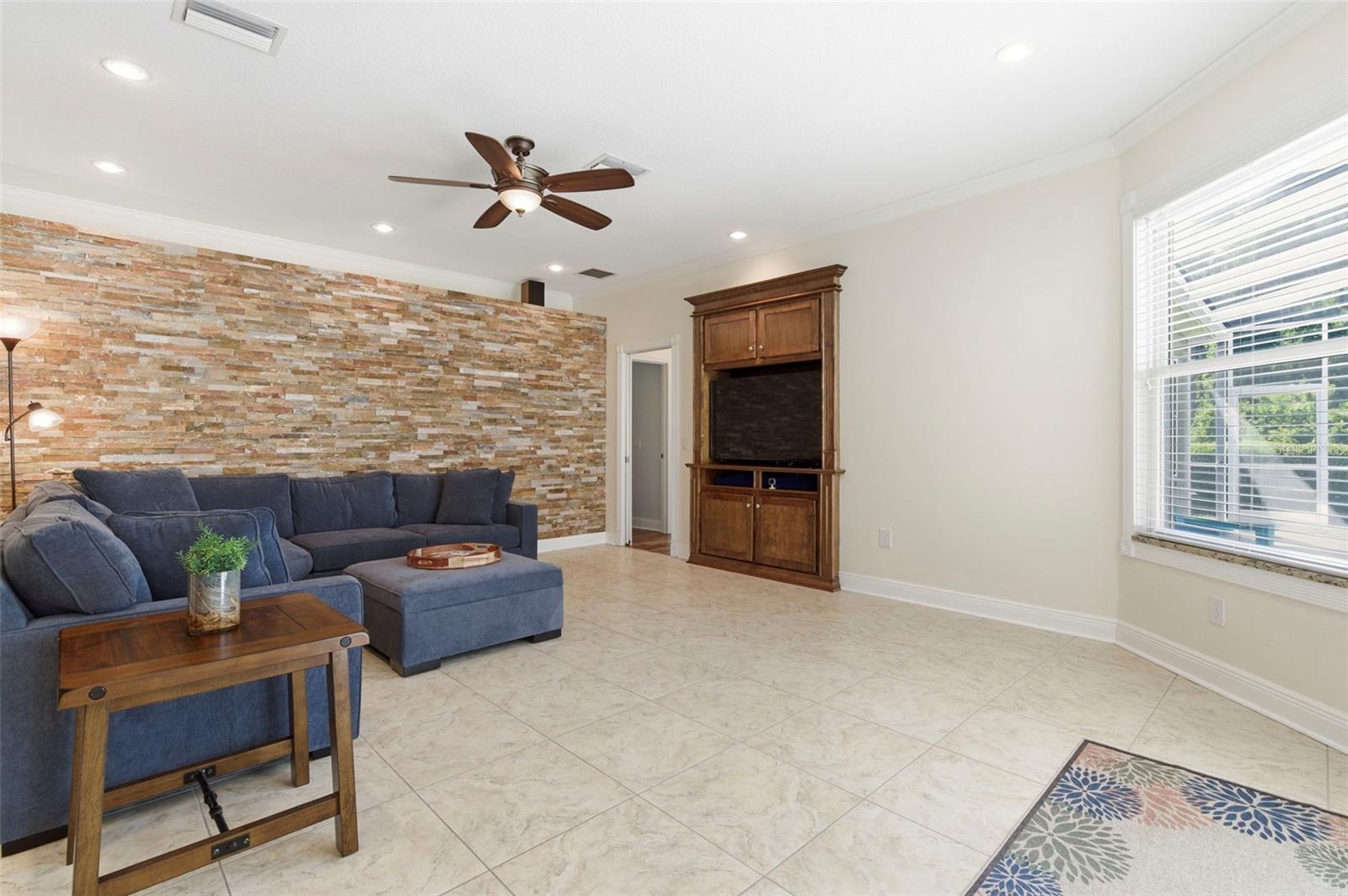 This spacious living area combines comfort and style with an open layout designed for both relaxing and entertaining. A striking stone accent wall creates a warm focal point, complemented by tile flooring, crown molding, and recessed lighting throughout. Natural light pours in through large windows, enhancing the bright and airy feel of the space. The room flows seamlessly into adjacent living areas, making it ideal for gatherings or everyday living.