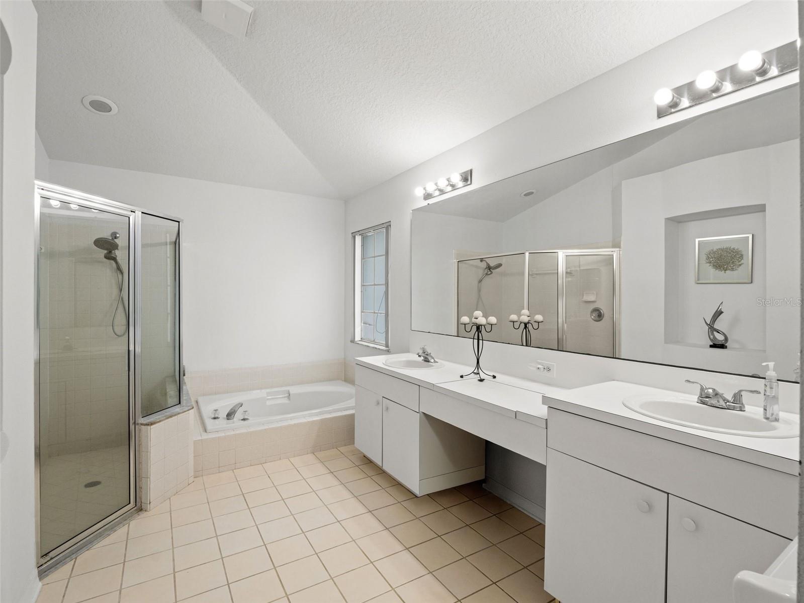 Large ensuite bath with dual vanities, expansive mirror, and a soaking tub alongside a separate glass-enclosed shower. Bright and open with plenty of counter space.