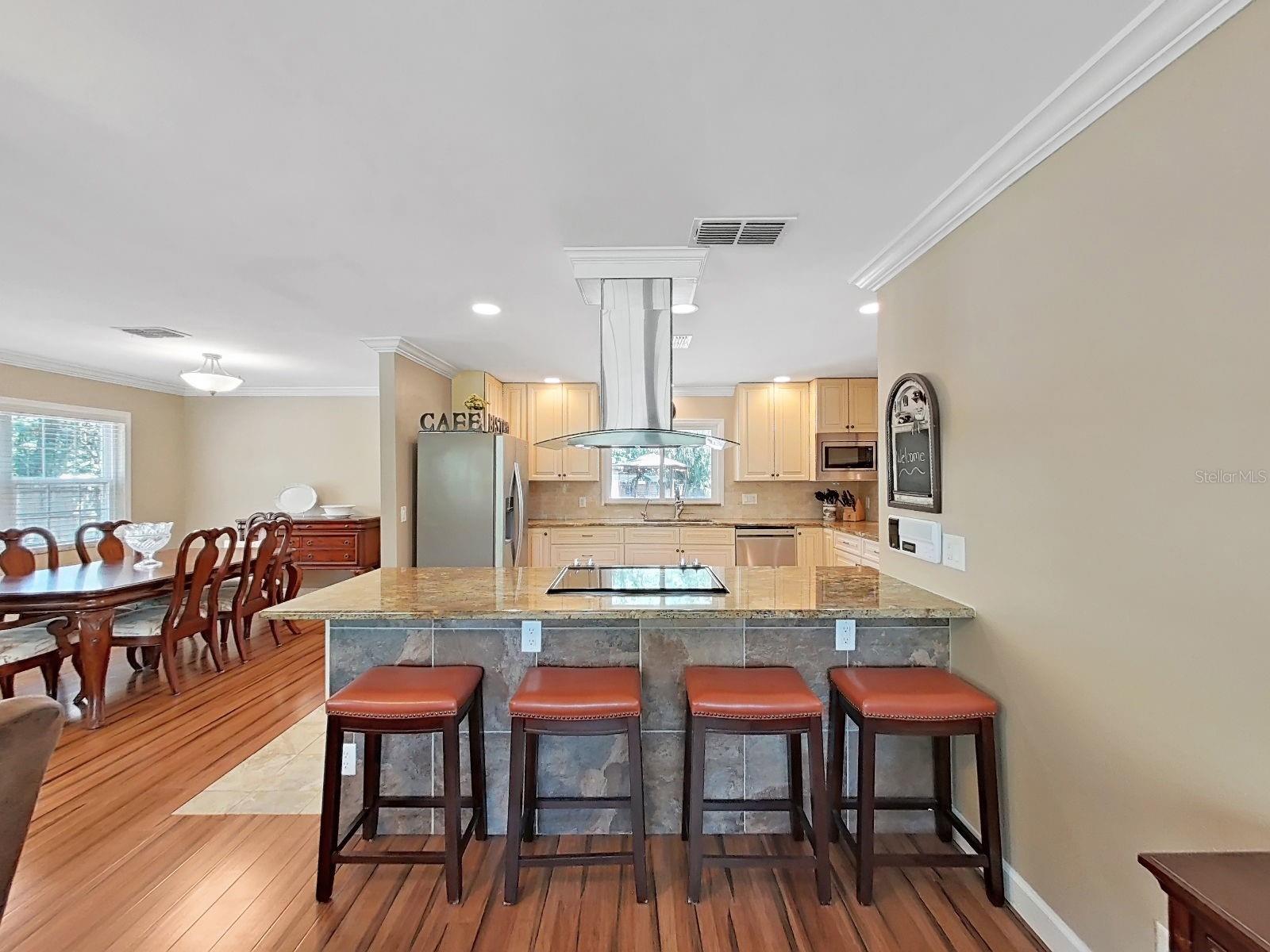 Stunning open concept kitchen featuring large granite peninsula with bar seating, sleek vent hood, & stainless appliances.