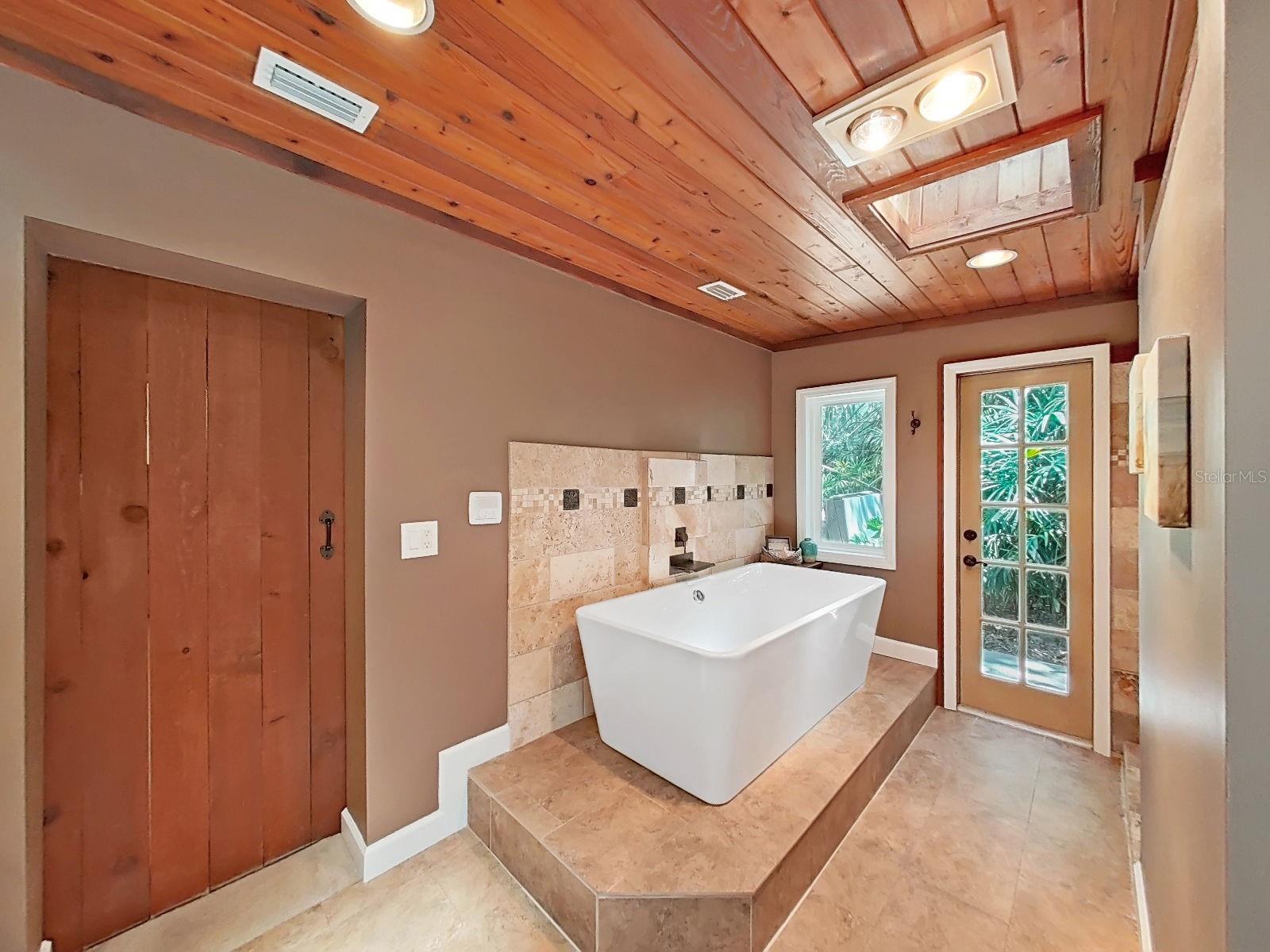 Spa-like primary bathroom with freestanding soaking tub on raised tile platform, wood-planked ceiling, skylight & french door with views - pure luxury!