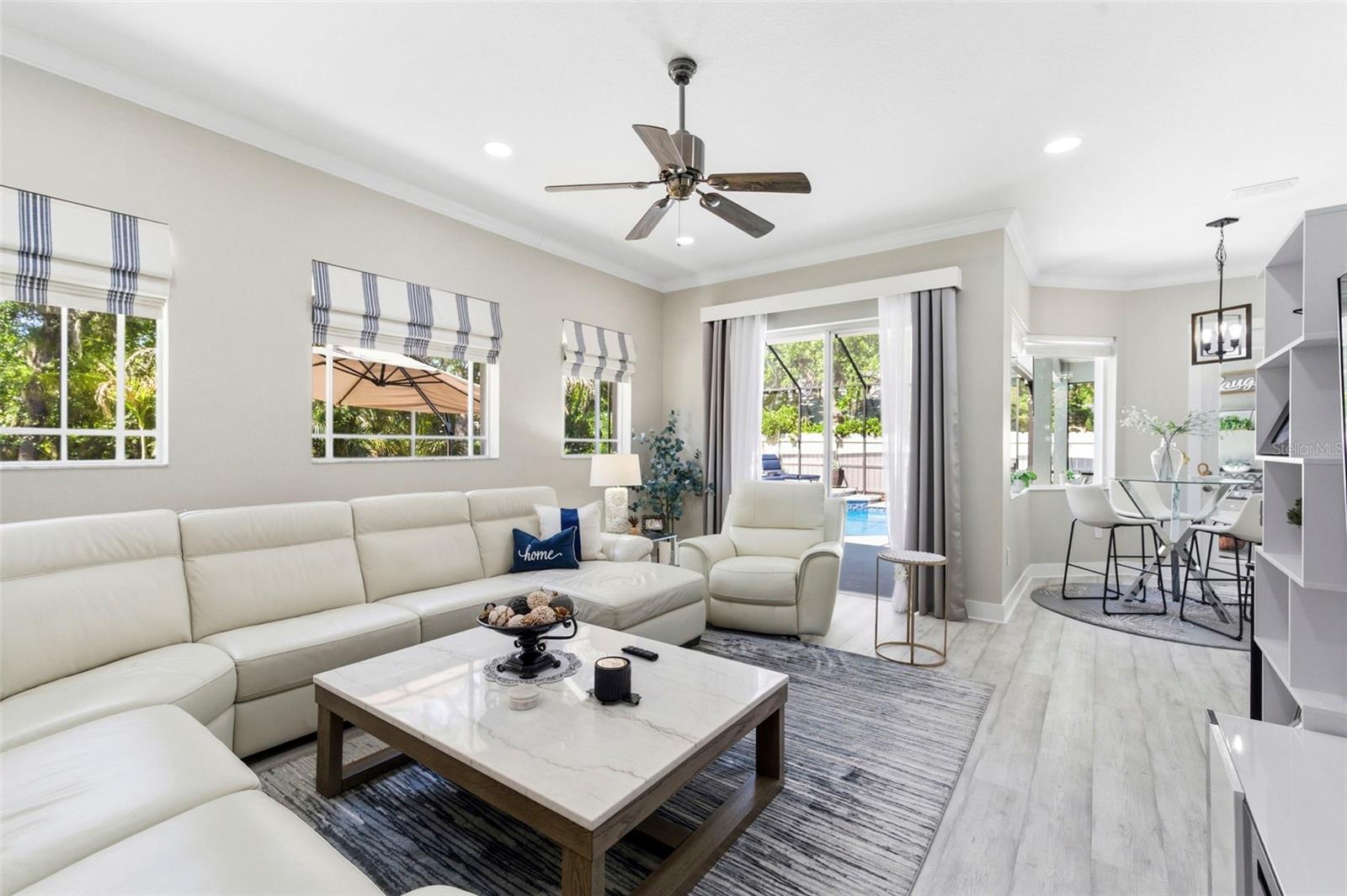 Bright and comfortable living room featuring a spacious sectional sofa, perfect for relaxing or entertaining. Large windows fill the space with natural light and offer pleasant views of the surrounding greenery. The room is complemented by neutral tones, modern finishes, and durable wood-look flooring, creating a clean and inviting atmosphere.