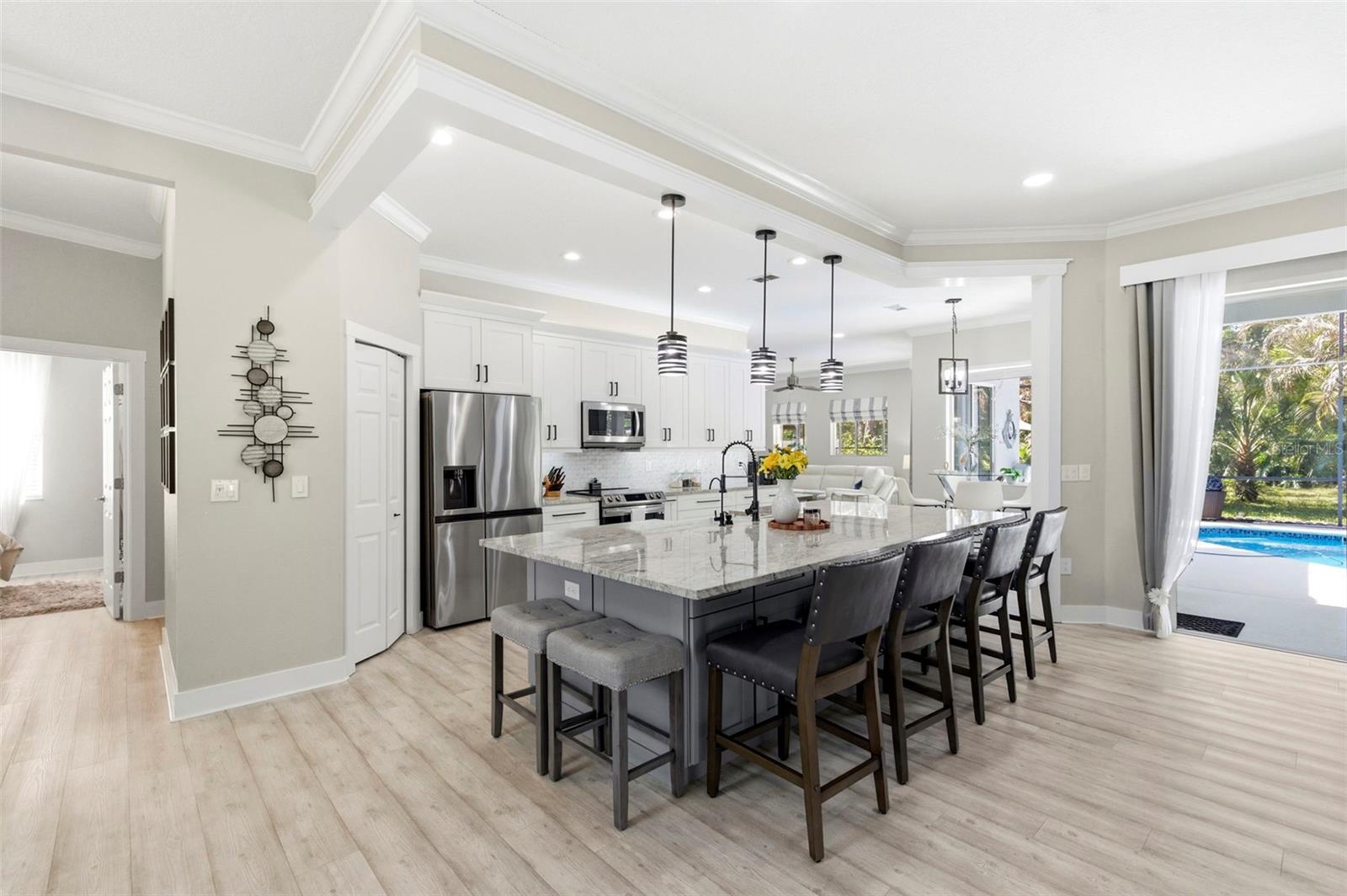 Beautifully designed open-concept living space showcasing a spacious chef’s kitchen with a large granite island, ample seating, and abundant cabinetry. Stainless steel appliances, modern pendant lighting, and elegant finishes create a clean, upscale feel. The layout flows seamlessly into the living and dining areas, with sliding glass doors opening to the pool area—perfect for indoor-outdoor entertaining and everyday living.