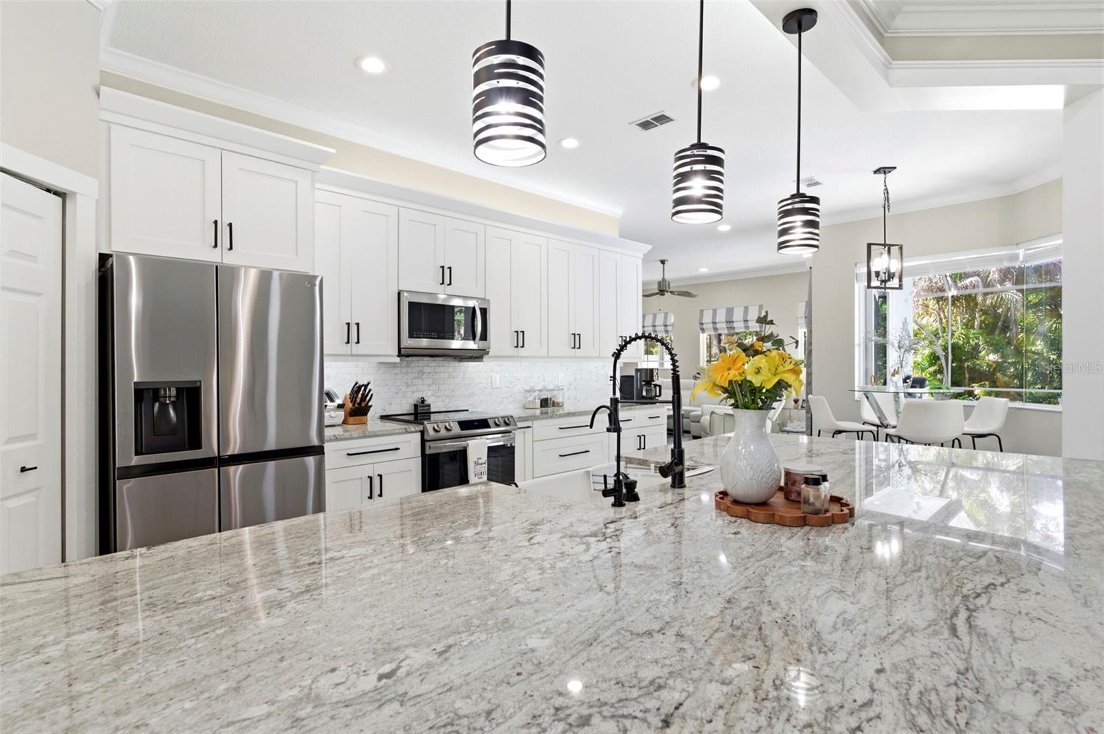 Stunning opBeautifully designed open-concept living space showcasing a spacious chef’s kitchen with a large granite island, ample seating, and abundant cabinetry. Stainless steel appliances, modern pendant lighting, and elegant finishes create a clean, upscale feel. The layout flows seamlessly into the living and dining areas, with sliding glass doors opening to the pool area—perfect for indoor-outdoor entertaining and everyday living.en-concept kitchen designed for both everyday living and entertaining, featuring a large center island with farmhouse sink, granite countertops, and ample cabinetry. Modern pendant lighting, stainless steel appliances, and sleek finishes create a clean, contemporary look. The space flows seamlessly into a bright dining area with large windows overlooking the pool and lush landscaping, while sliding glass doors provide easy indoor-outdoor living.