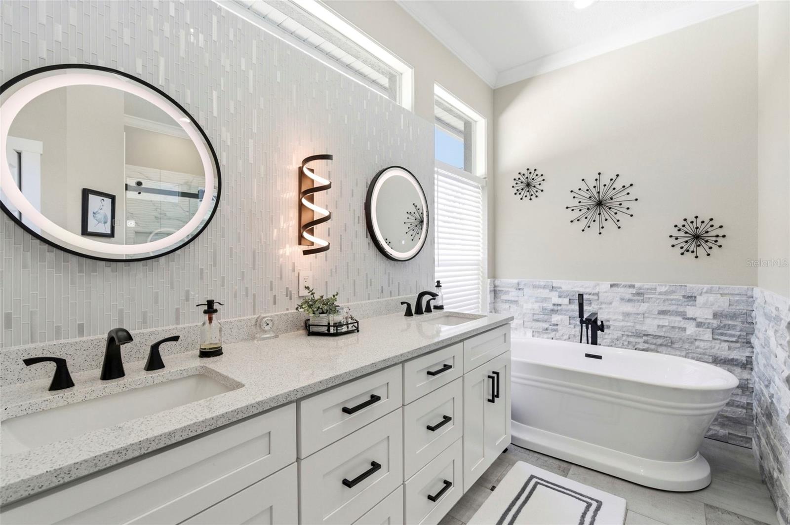 Spa-like primary bathroom featuring a dual-sink vanity with quartz countertops, shaker-style cabinetry, and matte black fixtures for a modern touch. A designer tile accent wall is complemented by elegant backlit mirrors and contemporary lighting. The space also offers a freestanding soaking tub with stylish tile surround, creating a relaxing retreat. Large windows provide abundant natural light, enhancing the bright, airy feel of this beautifully updated bathroom.