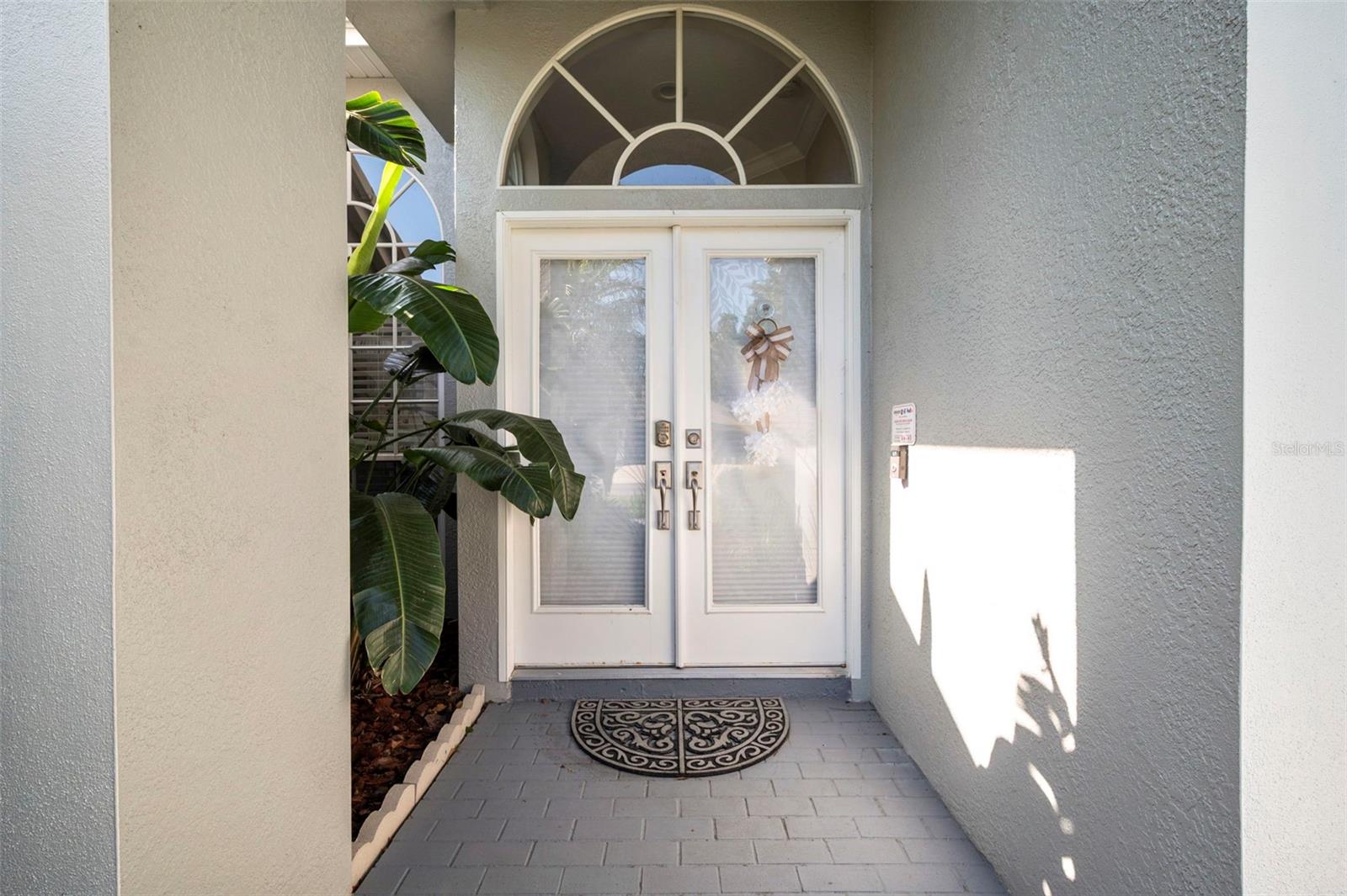 Inviting front entry featuring elegant double doors with decorative glass inserts and an arched transom window that adds architectural charm and natural light. The covered entryway offers a welcoming first impression, complemented by tropical landscaping and a clean, well-maintained exterior.