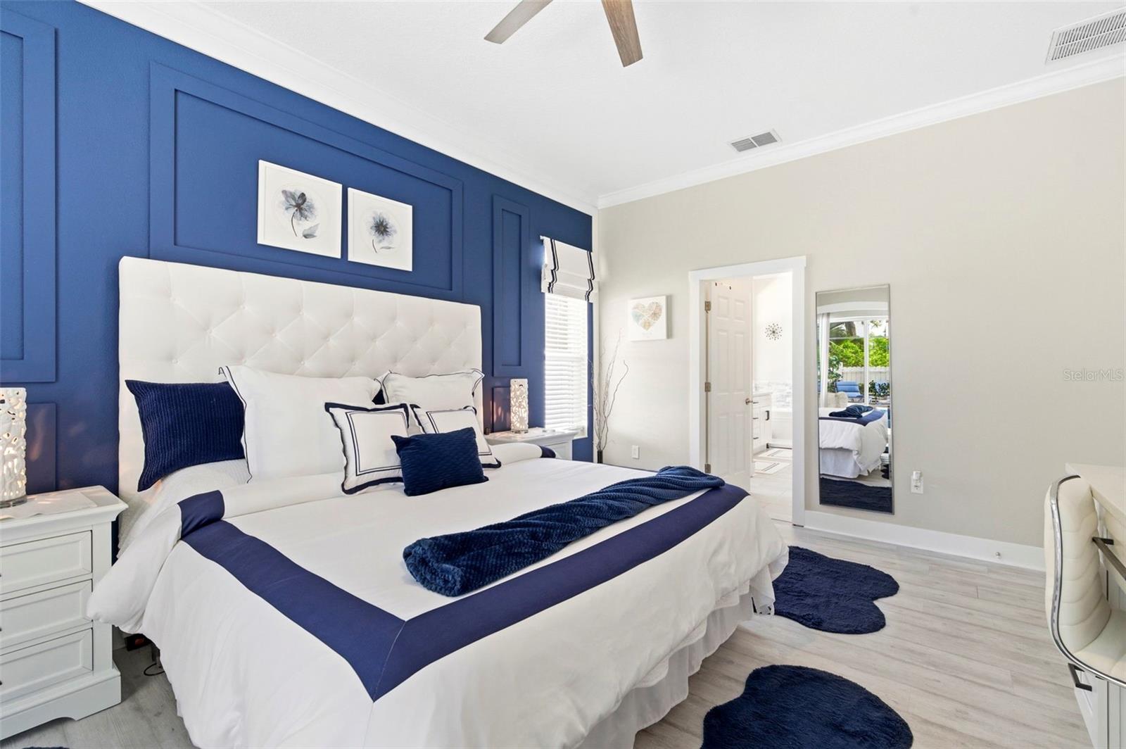 Stylish and serene primary bedroom featuring a striking accent wall with custom molding and designer finishes. The space offers a plush upholstered bed, coordinated décor, and a calming color palette that creates a true retreat. High ceilings and natural light enhance the open, airy feel, while the en-suite bathroom access adds convenience and functionality. A perfect blend of comfort and sophistication.