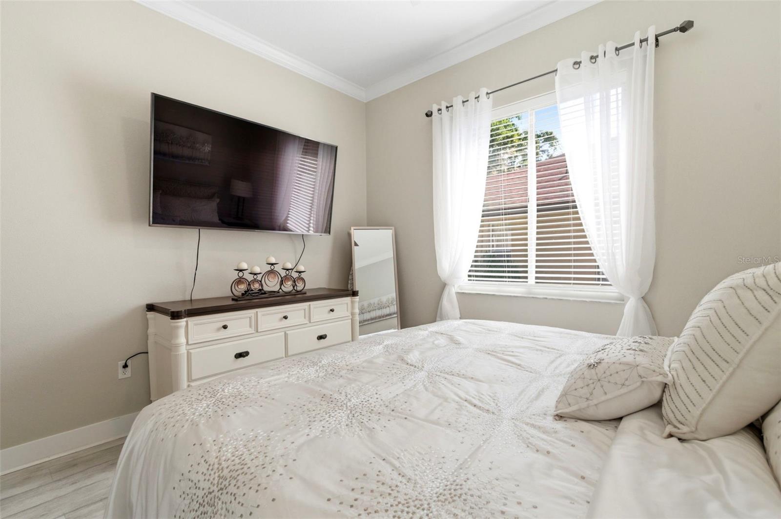 Light and airy bedroom featuring a soft neutral palette and elegant finishes throughout. Large windows with sheer curtains allow for abundant natural light, creating a warm and inviting atmosphere. The room offers a comfortable layout with stylish furnishings, ample space, and a serene feel—perfect for guests or family living.
