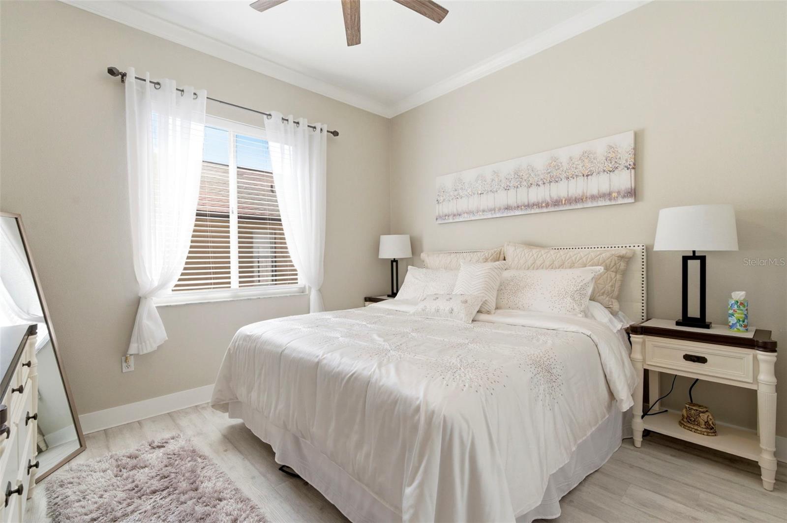 Light and airy bedroom featuring a soft neutral palette and elegant finishes throughout. Large windows with sheer curtains allow for abundant natural light, creating a warm and inviting atmosphere. The room offers a comfortable layout with stylish furnishings, ample space, and a serene feel—perfect for guests or family living.