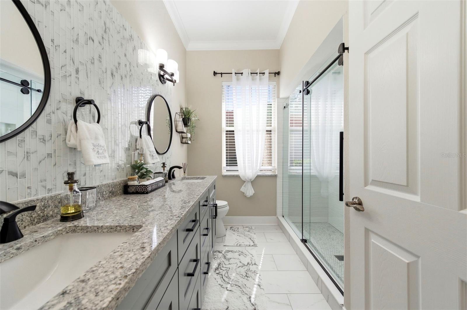 Beautifully updated bathroom featuring a dual-sink vanity with granite countertops, shaker-style cabinetry, and sleek matte black fixtures. A designer tile backsplash adds texture and elegance, while modern lighting and round mirrors enhance the contemporary feel. The spacious walk-in glass shower offers a clean, upscale look, and a large window brings in natural light, creating a bright and refreshing space.
