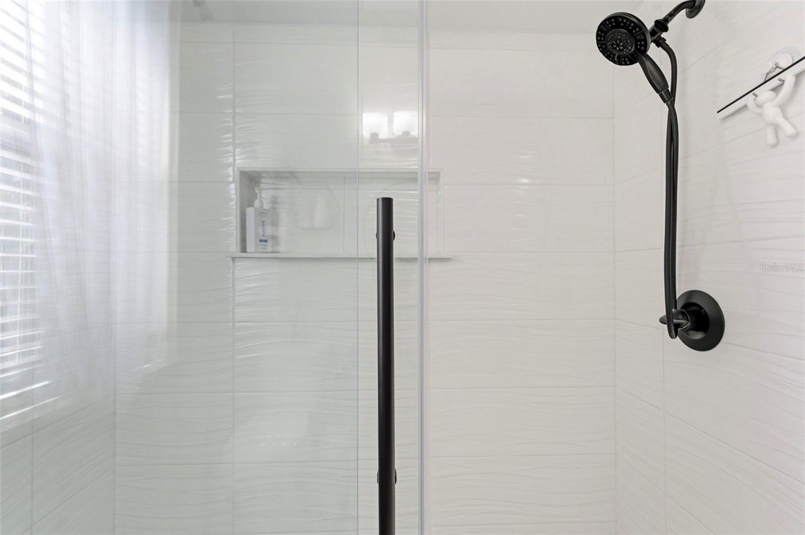 Beautifully updated bathroom featuring a dual-sink vanity with granite countertops, shaker-style cabinetry, and sleek matte black fixtures. A designer tile backsplash adds texture and elegance, while modern lighting and round mirrors enhance the contemporary feel. The spacious walk-in glass shower offers a clean, upscale look, and a large window brings in natural light, creating a bright and refreshing space.