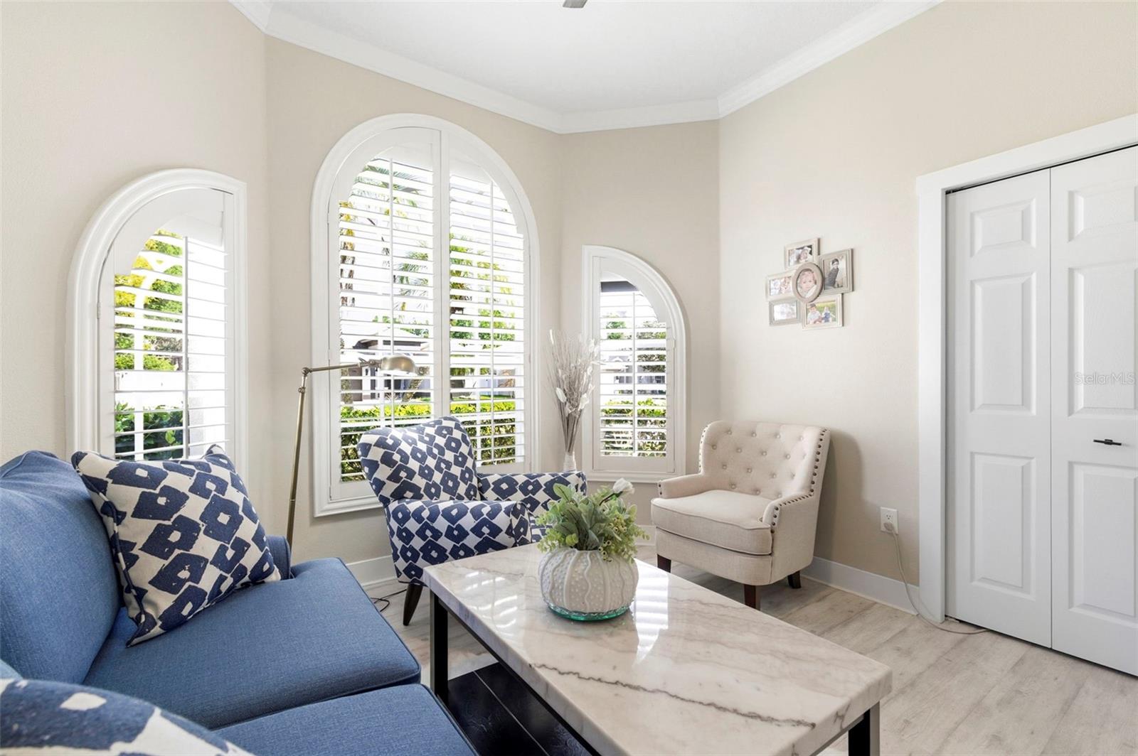 Bright and inviting living room featuring volume ceilings and abundant natural light. The space offers a comfortable layout with a plush sofa, accent chair, and a sleek marble-top coffee table, perfect for relaxing or entertaining. Neutral tones are complemented by tasteful décor, modern artwork, and a stylish shelving unit for added character and storage. Durable wood-look flooring completes the clean, contemporary feel of this cozy yet functional living area.