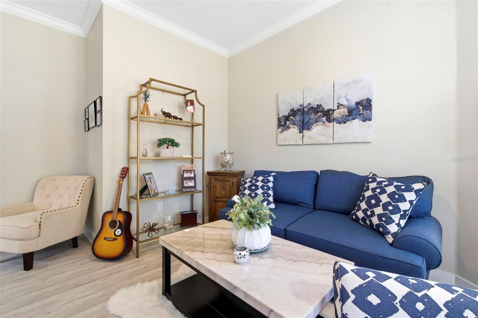 Bright and inviting living room featuring volume ceilings and abundant natural light. The space offers a comfortable layout with a plush sofa, accent chair, and a sleek marble-top coffee table, perfect for relaxing or entertaining. Neutral tones are complemented by tasteful décor, modern artwork, and a stylish shelving unit for added character and storage. Durable wood-look flooring completes the clean, contemporary feel of this cozy yet functional living area.