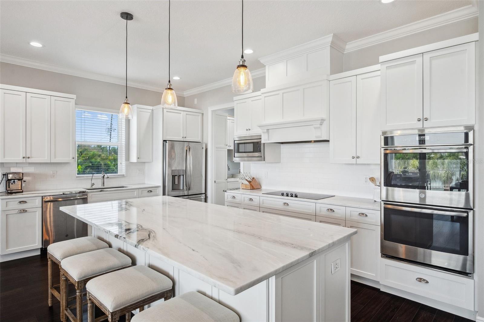 Gorgeous, marbled stone countertops and an oversized center island. Double ovens to the right and custom solid wood cabinetry and built ins throughout kitchen, living area and bar.