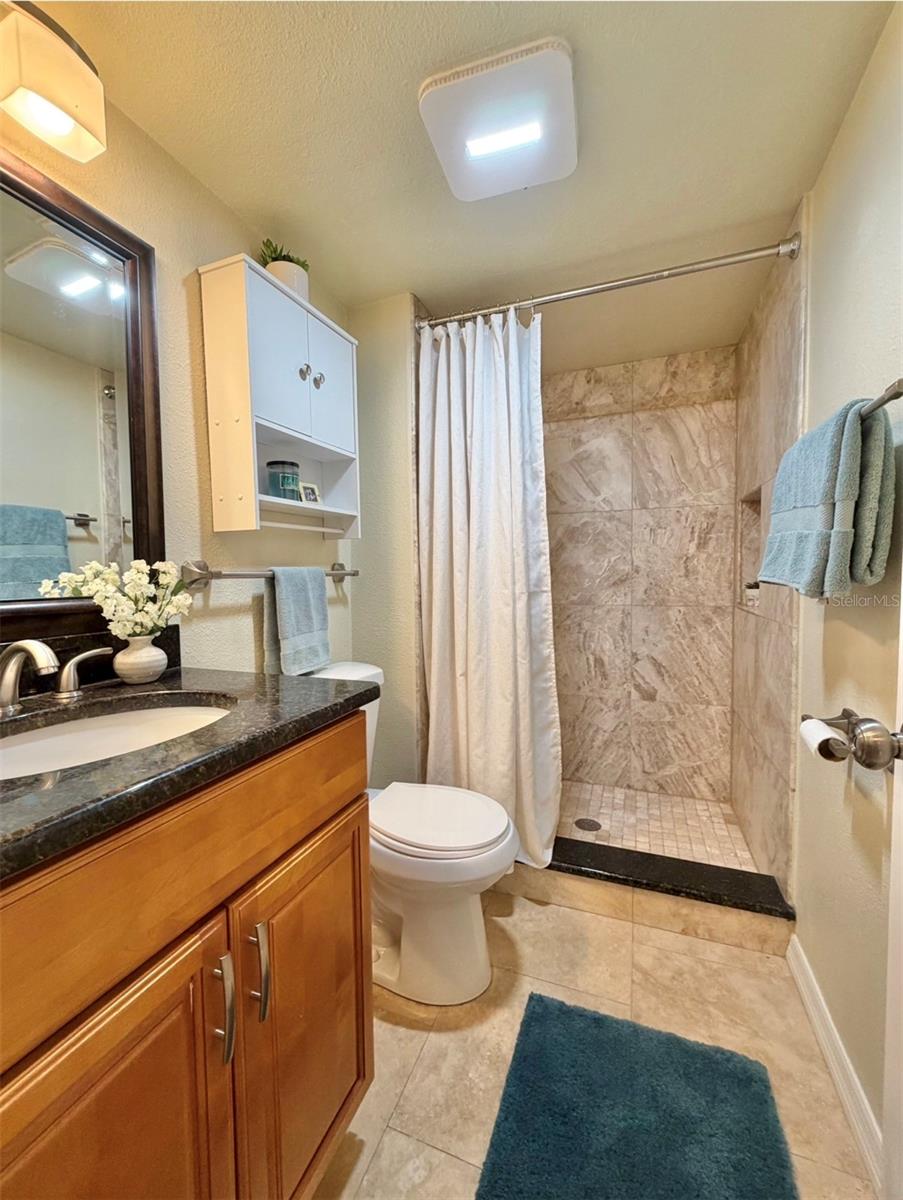 The second bathroom features a floor-to-ceiling tiled walk-in shower with a niche, granite countertop, a storage vanity, new satin nickel fixtures, a storage cabinet above the toilet, and updated lighting.