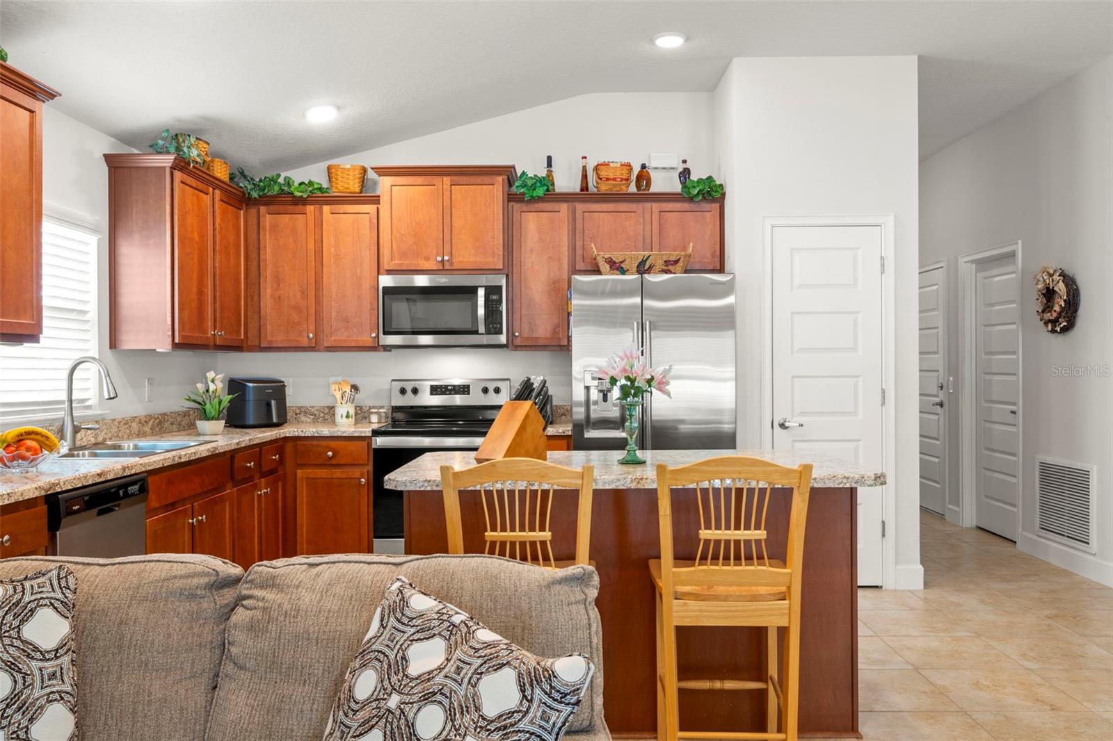 The open kitchen features warm wood cabinetry, stainless steel appliances, granite-look countertops, a center island with bar seating, dual sinks, a closet pantry and generous prep and storage space.