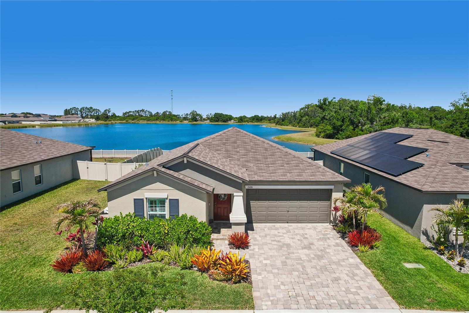 Elevated view of the home's stucco exterior, pavered driveway, and tropical landscaping with a large pond directly behind the property. Mature tree line frames the water on the far shore.