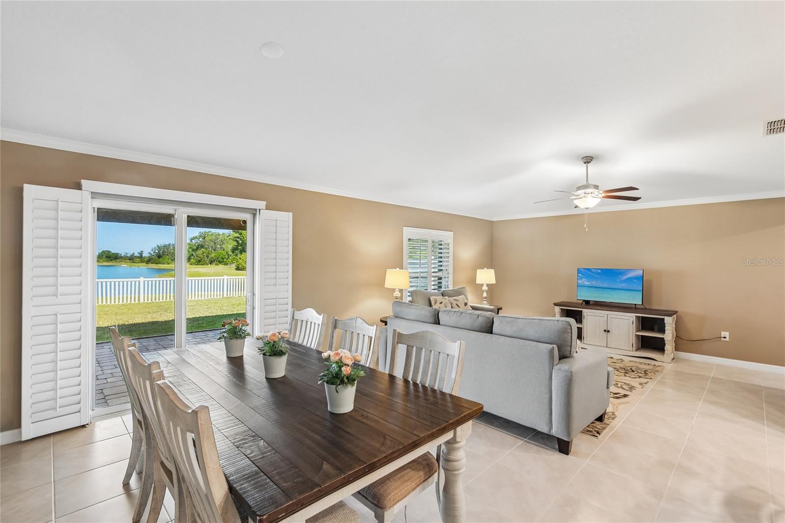 Dining area and great room with tile flooring, crown molding, ceiling fan, and plantation shutters. Sliding glass doors open to a pavered patio with a direct pond view beyond the rear fence line.