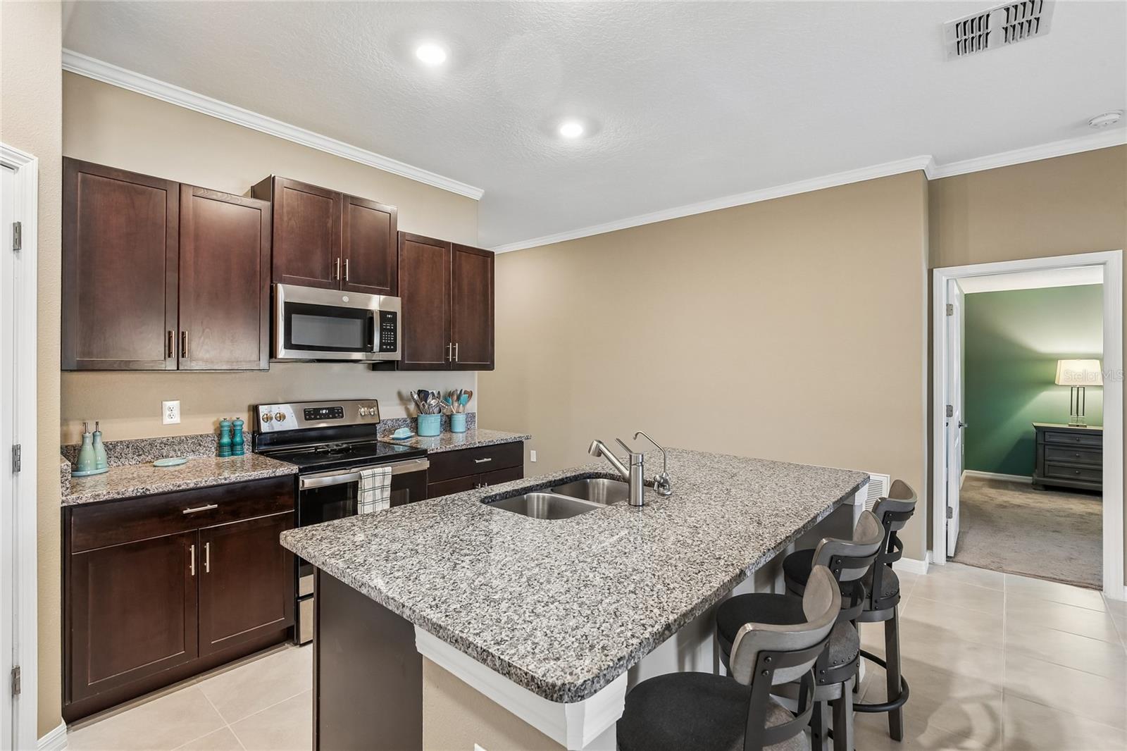 Kitchen with espresso cabinetry, granite countertops, stainless steel range and built-in microwave, and a peninsula island with an undermount sink and barstool seating. Tile flooring and crown molding carry through from the main living areas.