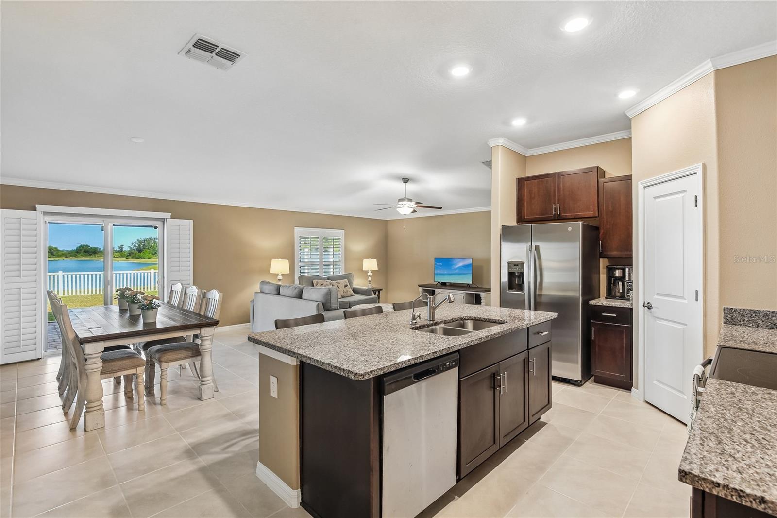 Kitchen island with granite countertops, undermount sink, and dishwasher anchors an open floor plan extending to the dining area, great room, and sliding glass doors with a pond view. Stainless steel French door refrigerator, espresso cabinetry, recessed lighting, and crown molding throughout.