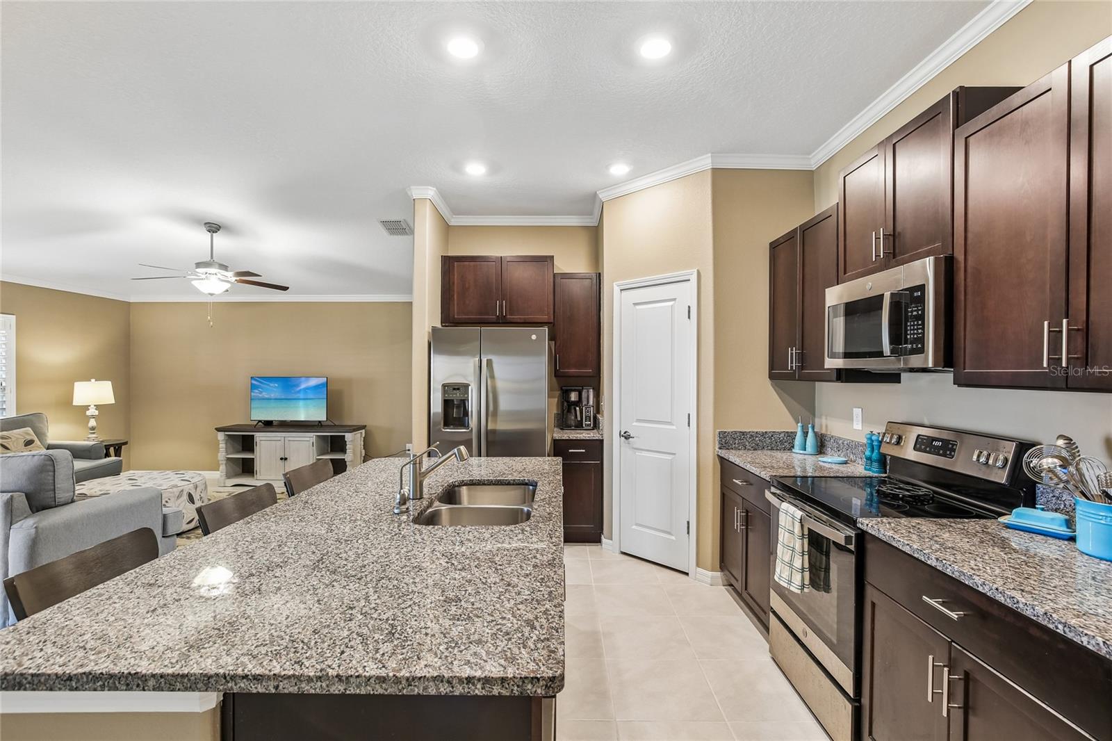 Kitchen with dark espresso cabinetry, granite countertops, stainless steel appliances including a French door refrigerator and built-in microwave, and a large island with an undermount sink and breakfast bar seating. Crown molding and recessed lighting throughout; open sightlines to the great room.