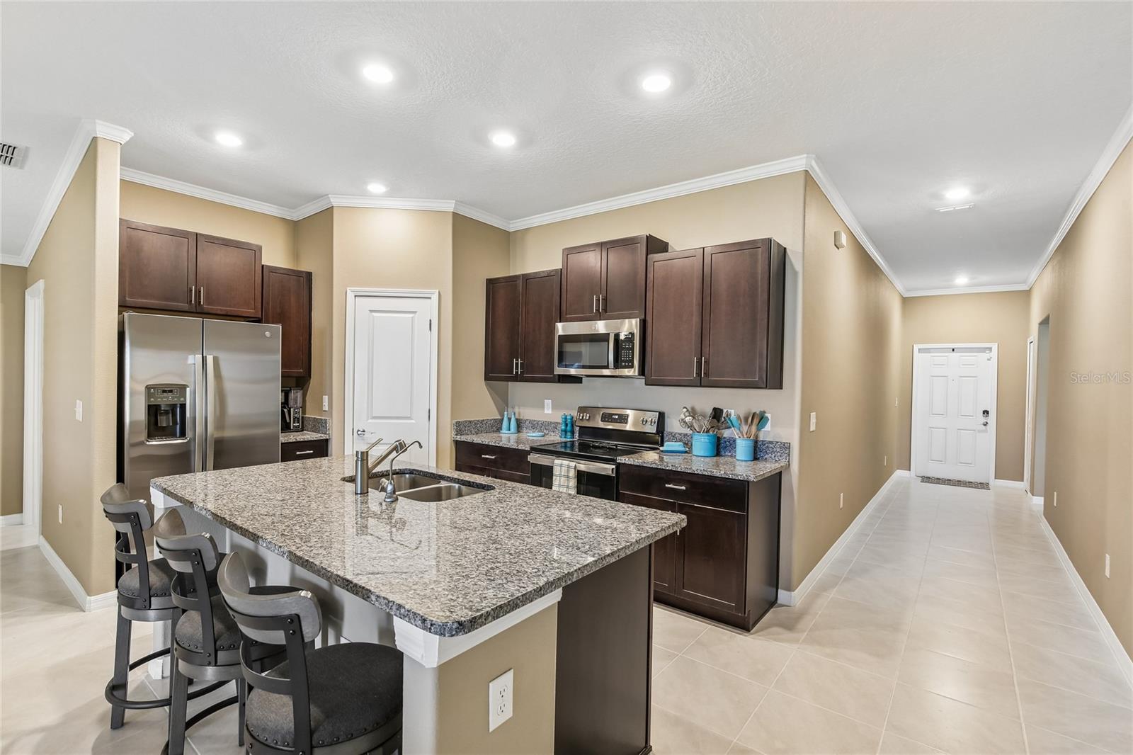 Kitchen with espresso cabinetry, granite countertops, stainless steel appliances including a French door refrigerator, electric range, and built-in microwave, and a center island with an undermount sink and barstool seating. Recessed lighting and crown molding throughout; tile flooring extends into the adjacent hallway.