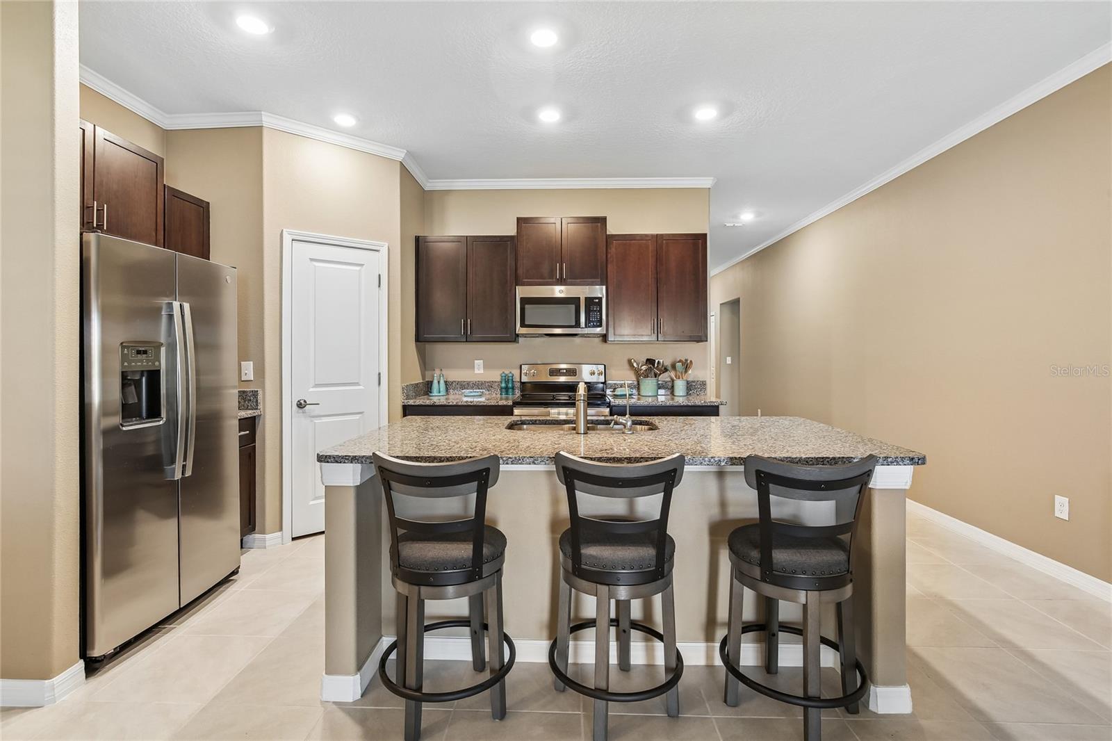 Kitchen island with granite countertops, undermount sink, and three-seat breakfast bar faces the cooking area with espresso cabinetry, stainless steel range and built-in microwave, and a French door refrigerator. Recessed lighting and crown molding carry through the space.